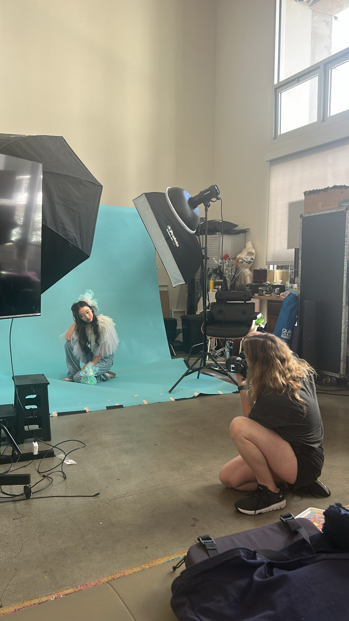 Person sitting on the floor in front of a photo shoot setup with a model on a blue background, studio equipment visible.