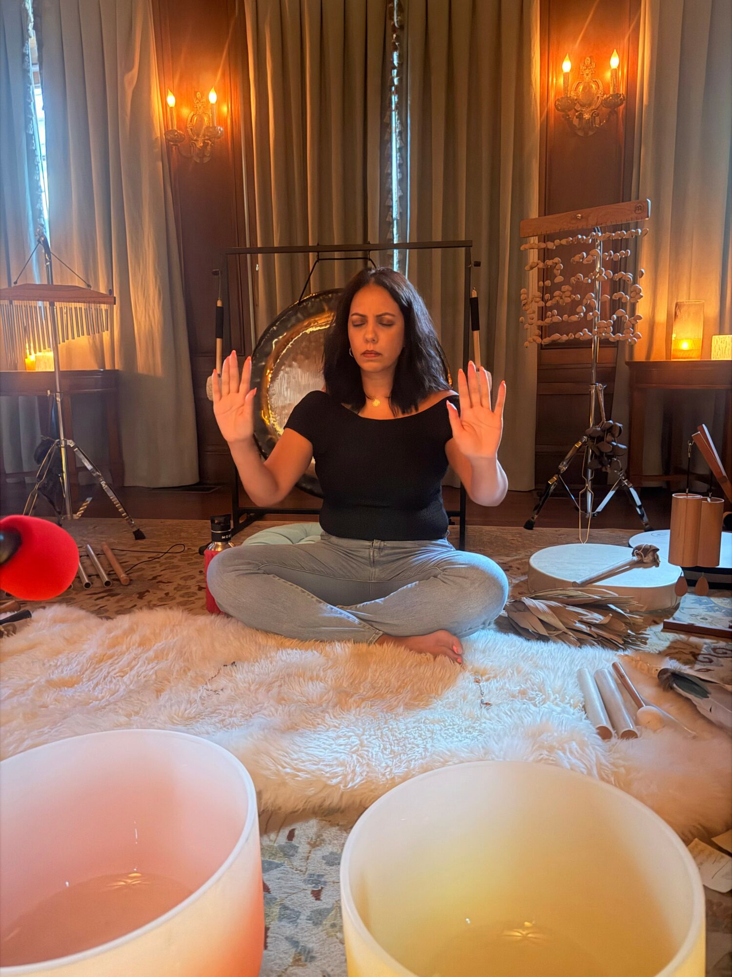 Woman sitting cross-legged on a rug with eyes closed and hands raised, surrounded by musical instruments and candles in a dimly lit room.