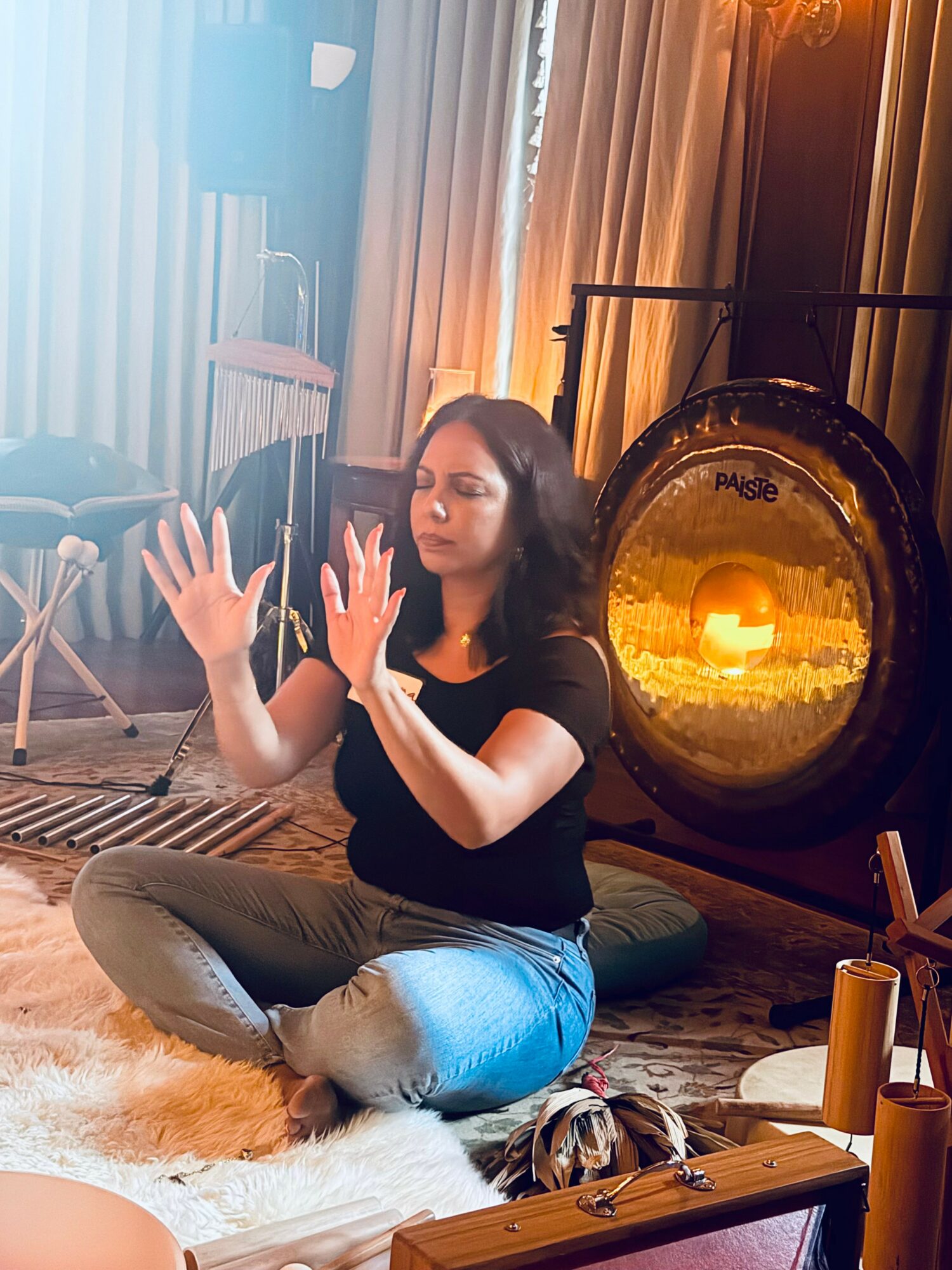 Woman sitting cross-legged on a rug with hands raised, in a room with curtains and a gong in the background.
