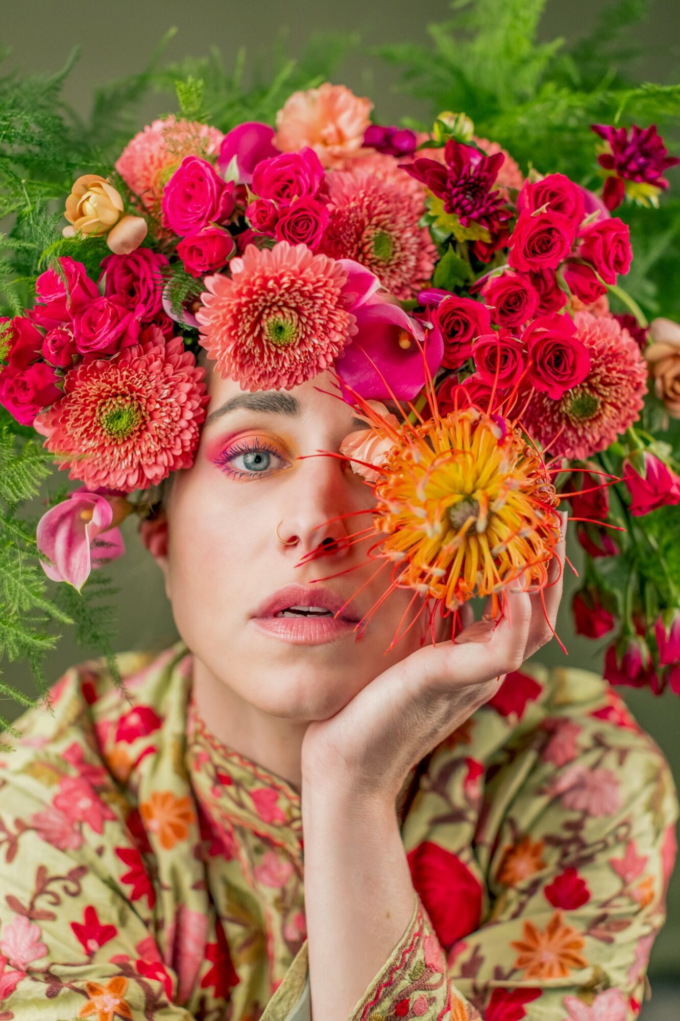 Woman with floral headpiece and colorful makeup, holding a flower near her face, against a green background.