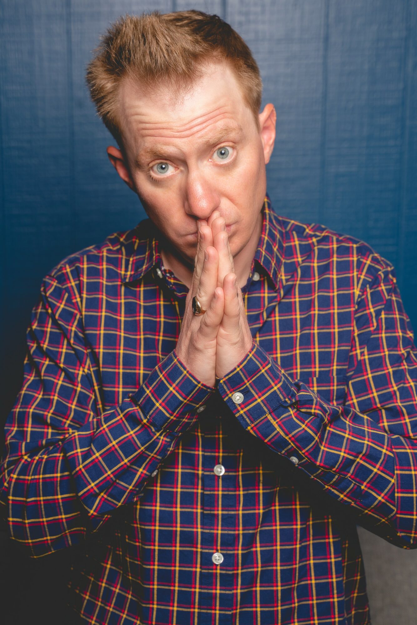 Man with short hair and plaid shirt holds hands near face, looking surprised, against a blue background.