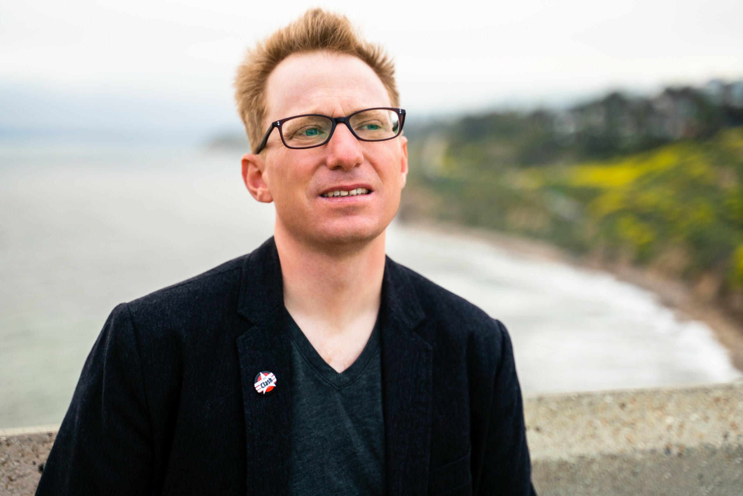 Man with glasses and short hair standing outdoors near a body of water, with a blurred landscape in the background.