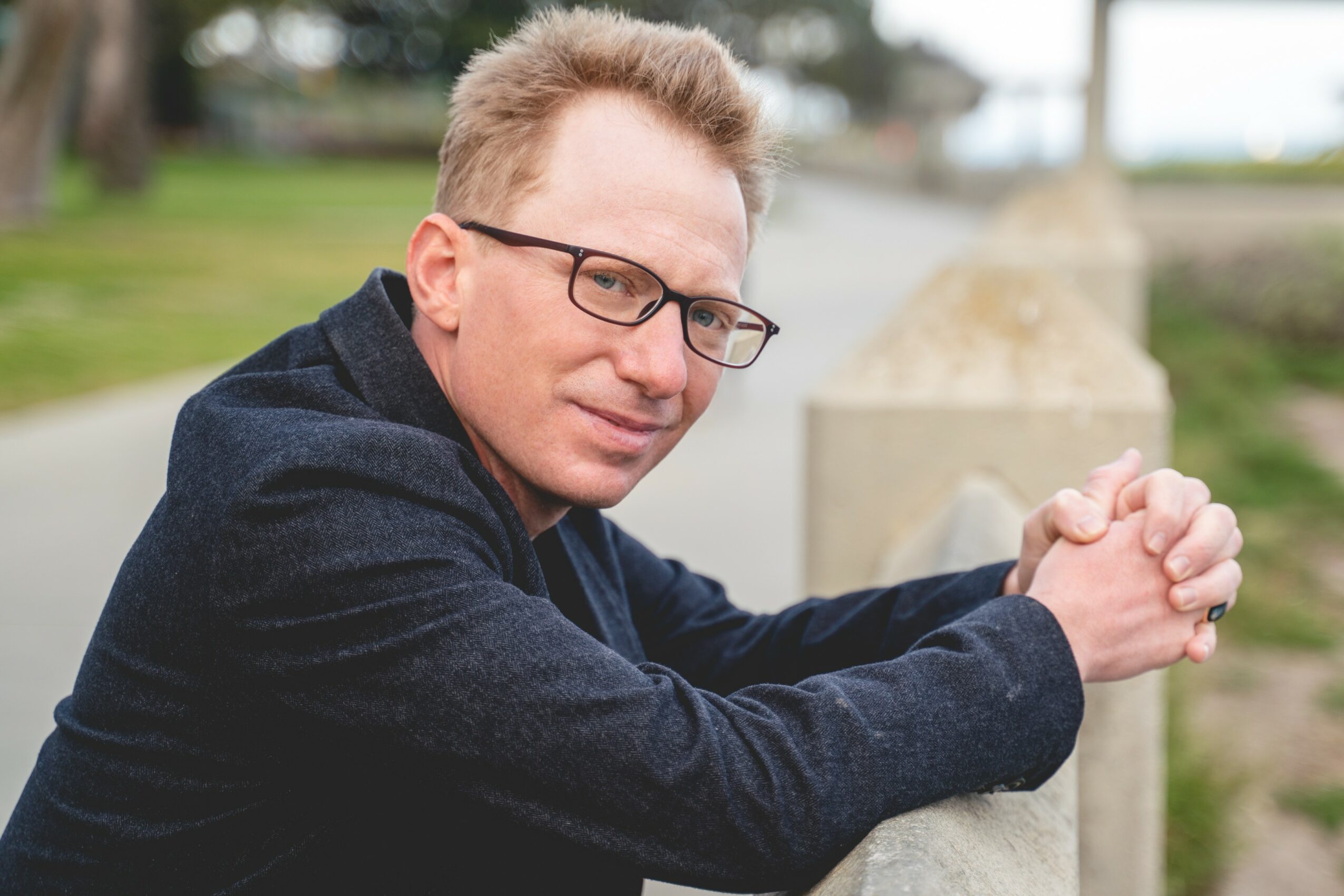 Man with glasses and short hair leaning on a concrete barrier outdoors, smiling, with trees and a pathway in the background.