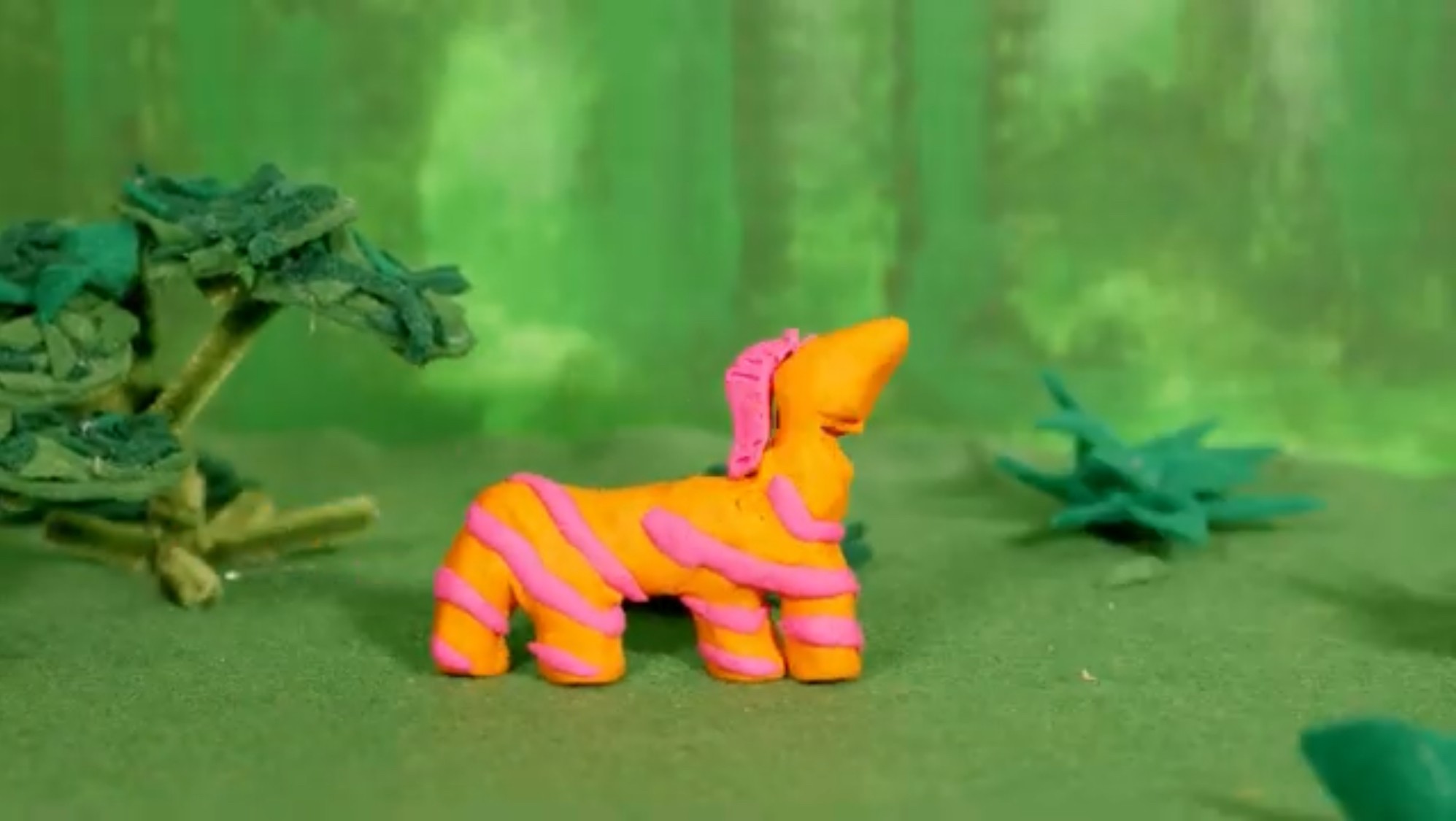 Colorful plush toy resembling a striped animal with a pink mane, set in a green forest background.