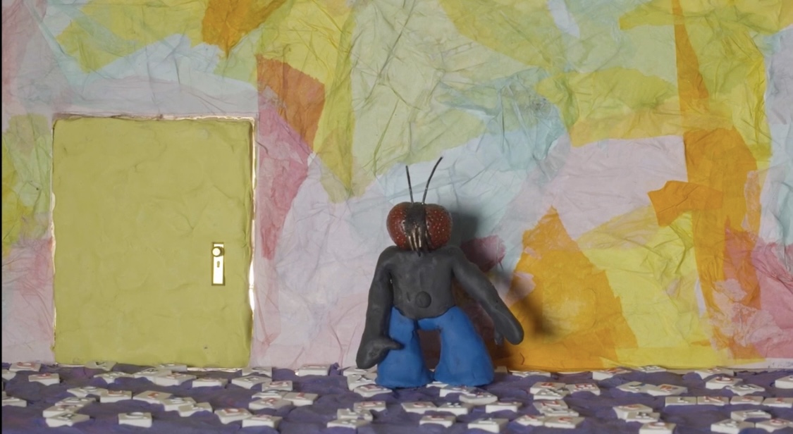 Small puppet with a red head and blue pants sits on a patterned surface in front of a colorful, abstract background.