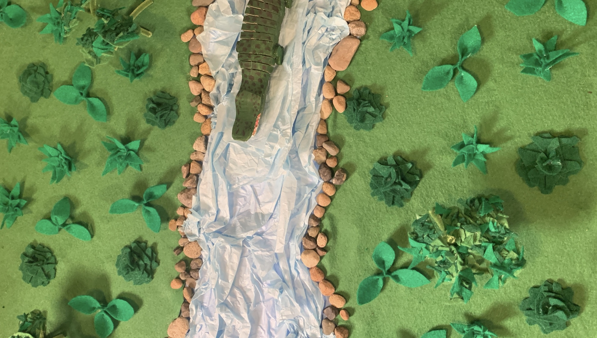 Paper craft landscape with a river, trees, and plants, viewed from above, with a green background.