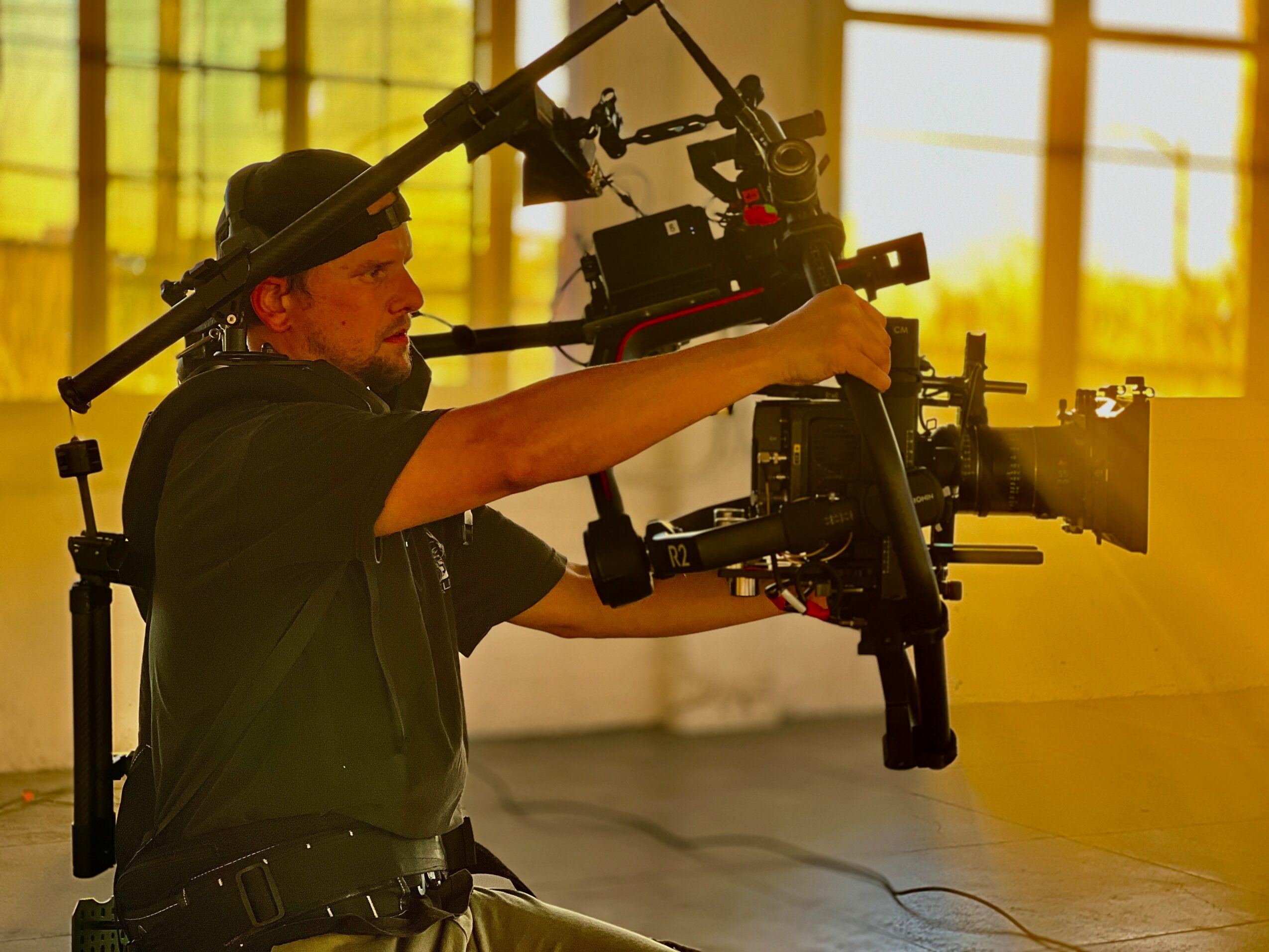Person operating a camera rig indoors with sunlight streaming through windows, wearing a black cap and shirt.