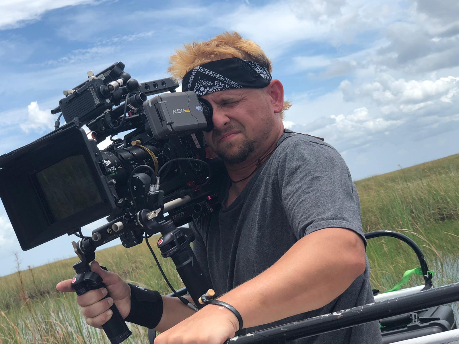 Man with a camera outdoors in a grassy field, wearing a bandana and gray t-shirt, holding a professional camera setup.