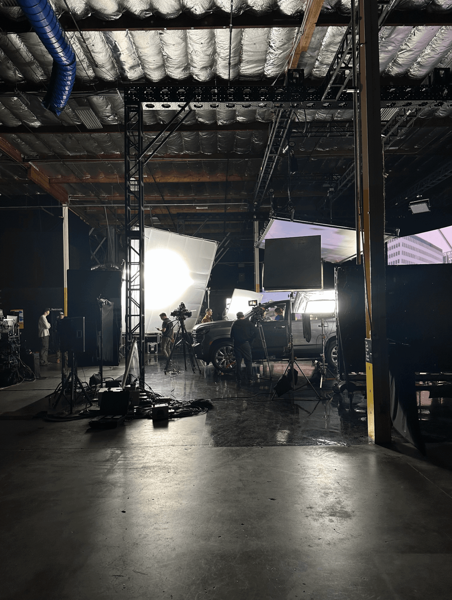 Studio or warehouse with lighting equipment, vehicles, and people, viewed from a low angle, with bright light in the background.