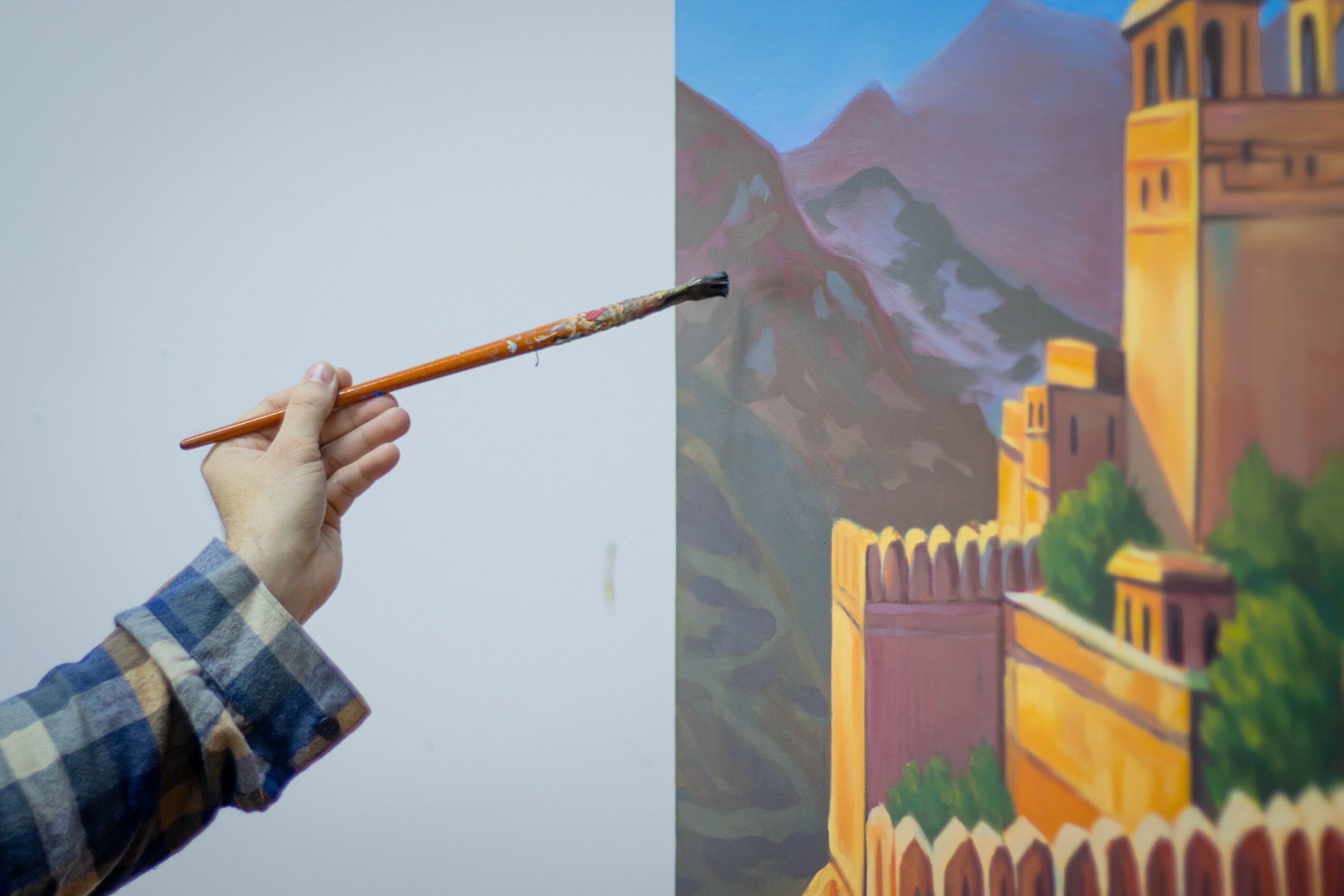 A person holds a paintbrush in front of a painted landscape of a castle on a mountain, with colorful walls and towers.