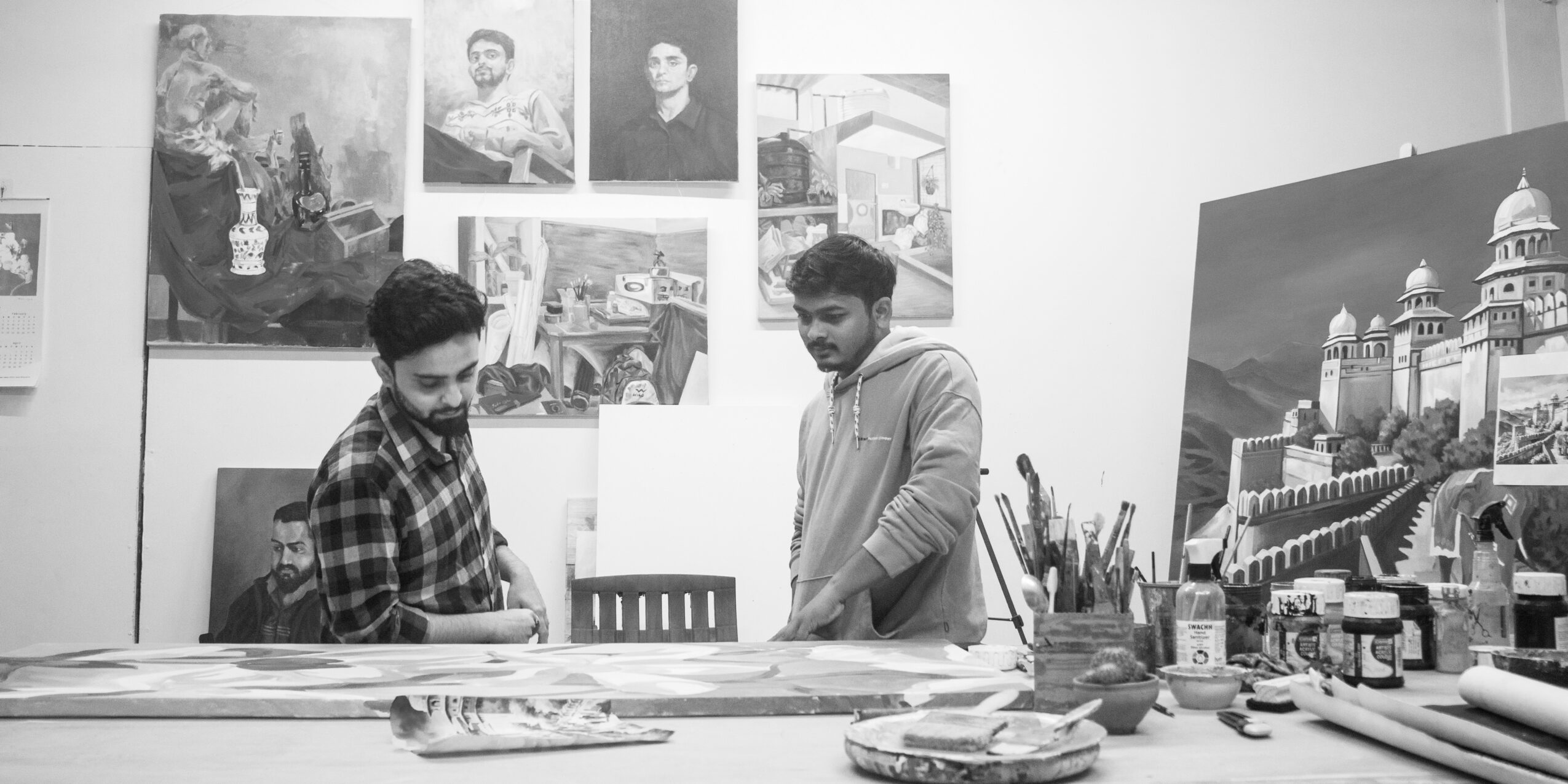 Two people in an art studio looking at paintings and art supplies on a table, with artwork on the walls behind them.
