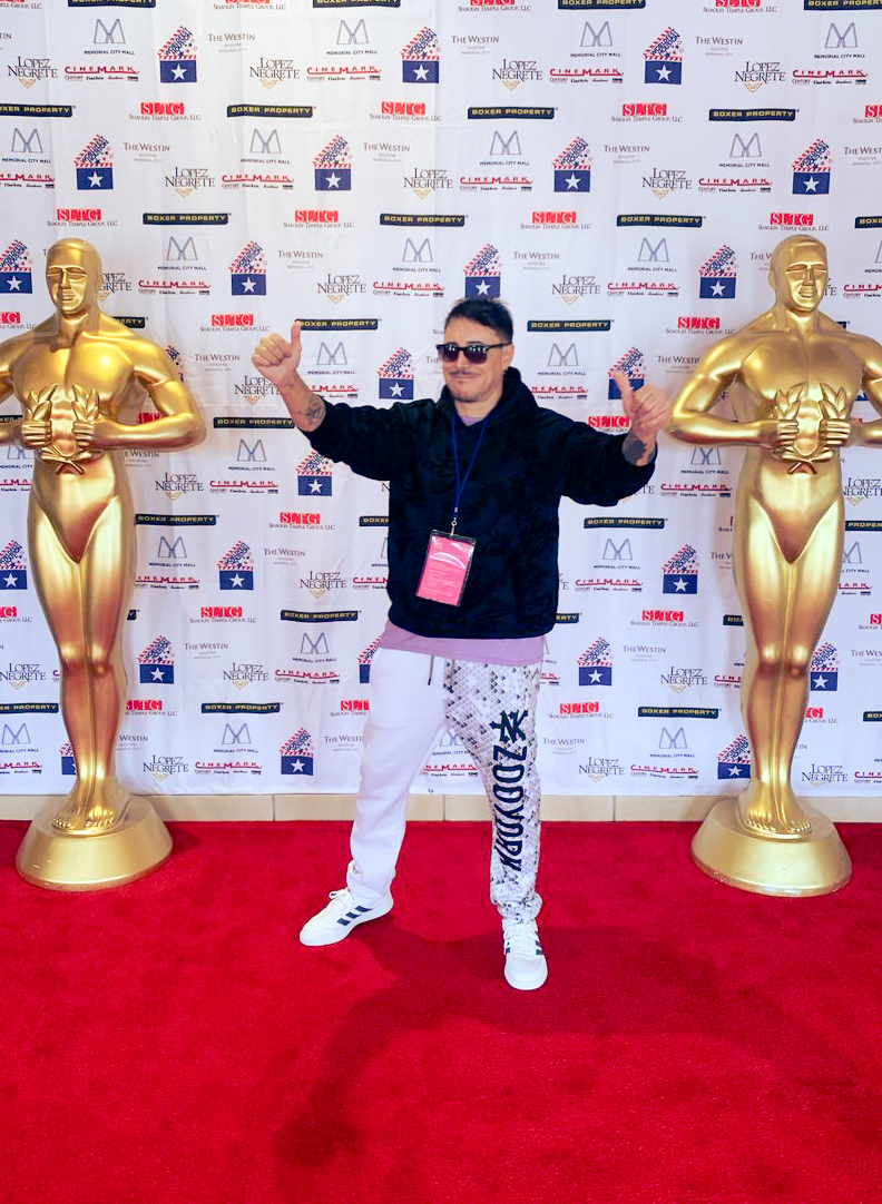 Person standing on red carpet between two large gold statues, holding a trophy, wearing sunglasses, black jacket, and white pants.