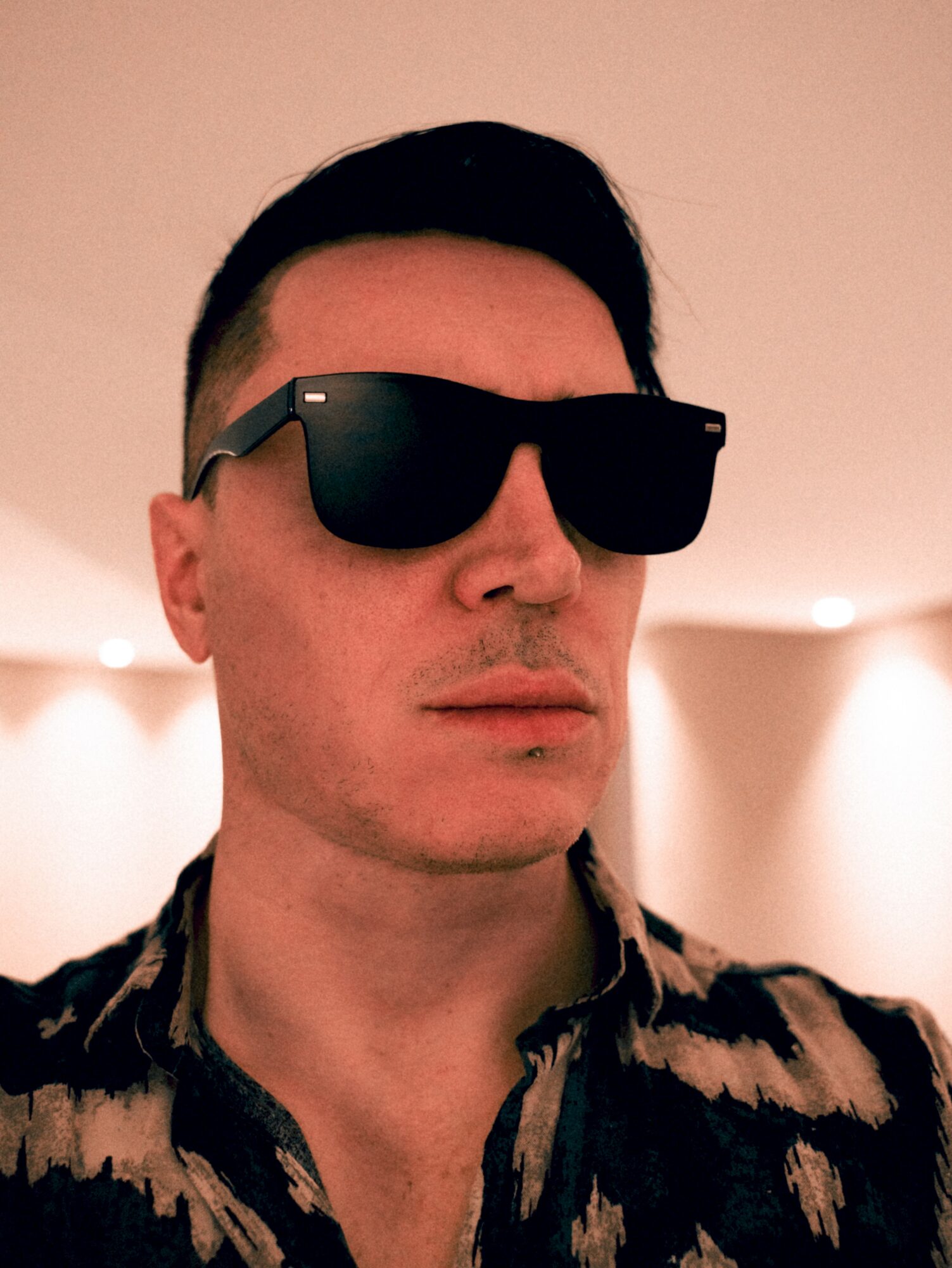 Man wearing sunglasses and patterned shirt, facing forward, indoors with soft lighting.