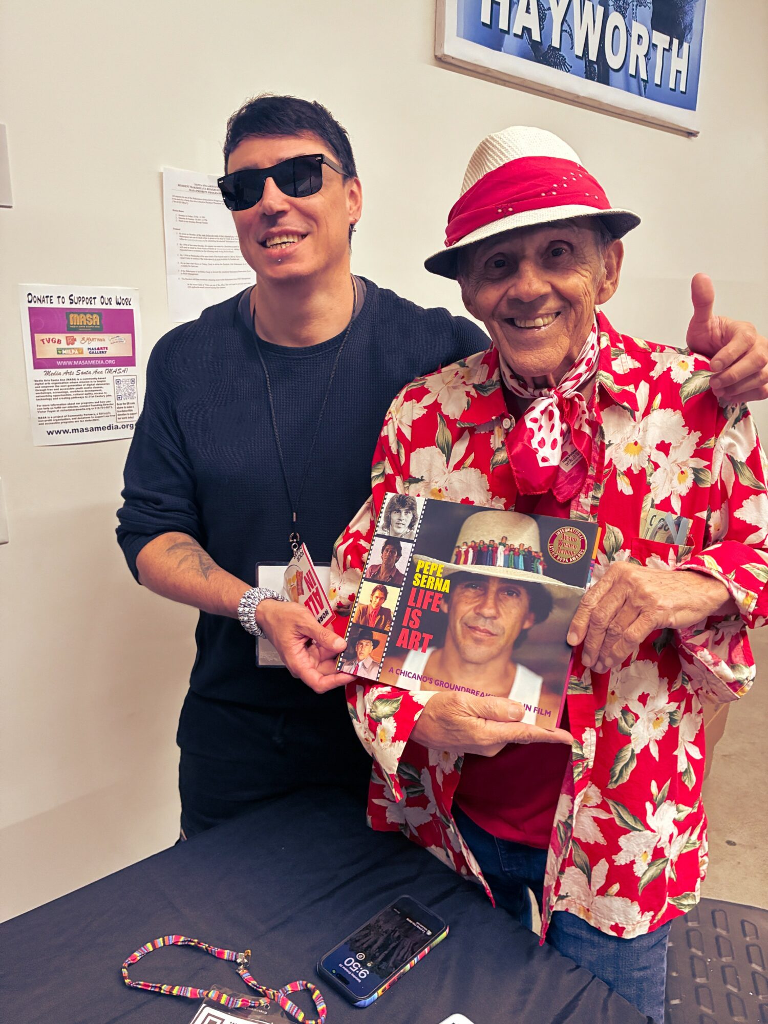 Two men smiling, one wearing sunglasses and a dark shirt, the other in a red floral shirt and hat, holding a magazine.