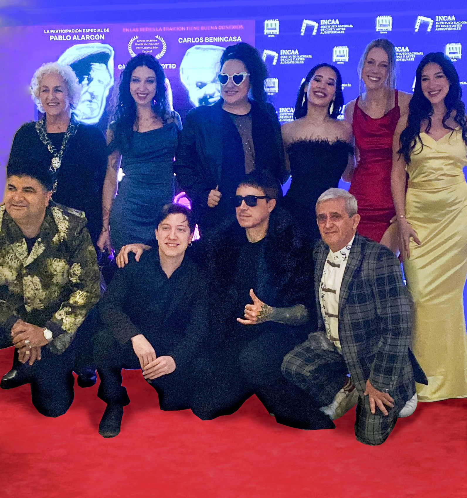 Group of ten people posing on red carpet, some standing and some kneeling, with a blue backdrop and screen behind them.