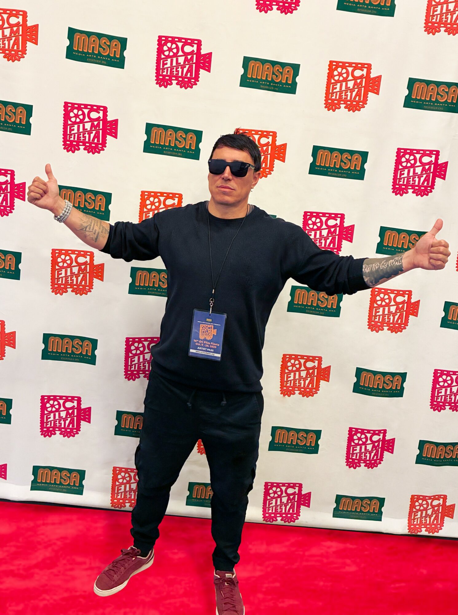 Man standing with arms outstretched in front of a patterned backdrop, wearing sunglasses and a black outfit.