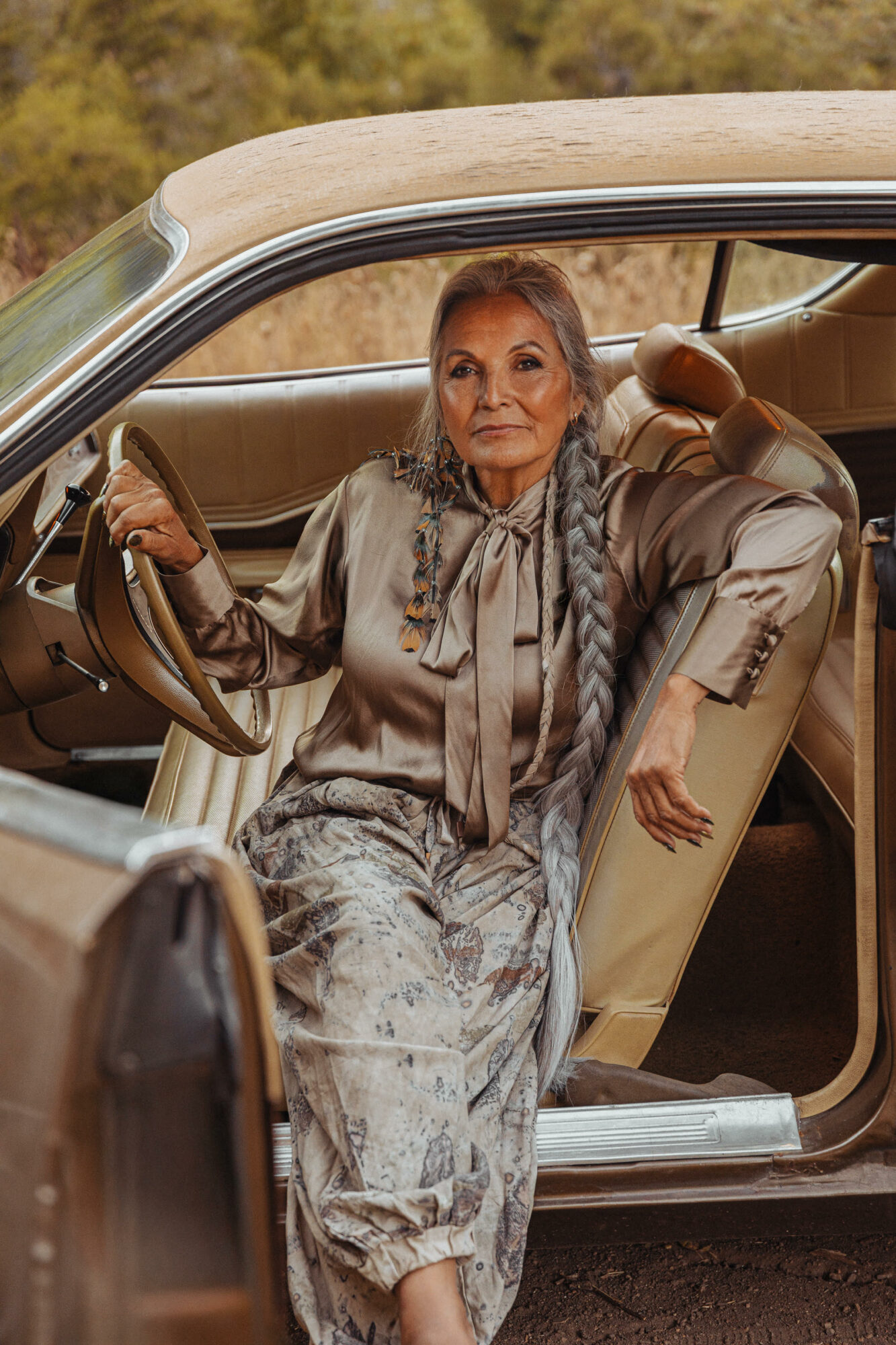 Elderly woman with long braided hair sitting in a vintage car, outdoors with trees in the background.