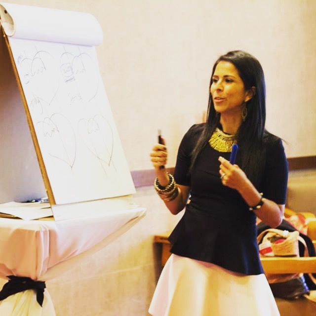 Woman standing in front of a flip chart with sketches, holding a marker, wearing a black top and white skirt, in a room.