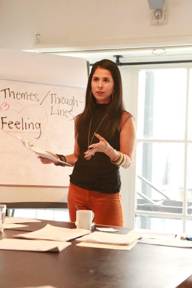 Woman standing in front of a whiteboard, holding a marker and gesturing, with papers on a table in front, near a window.
