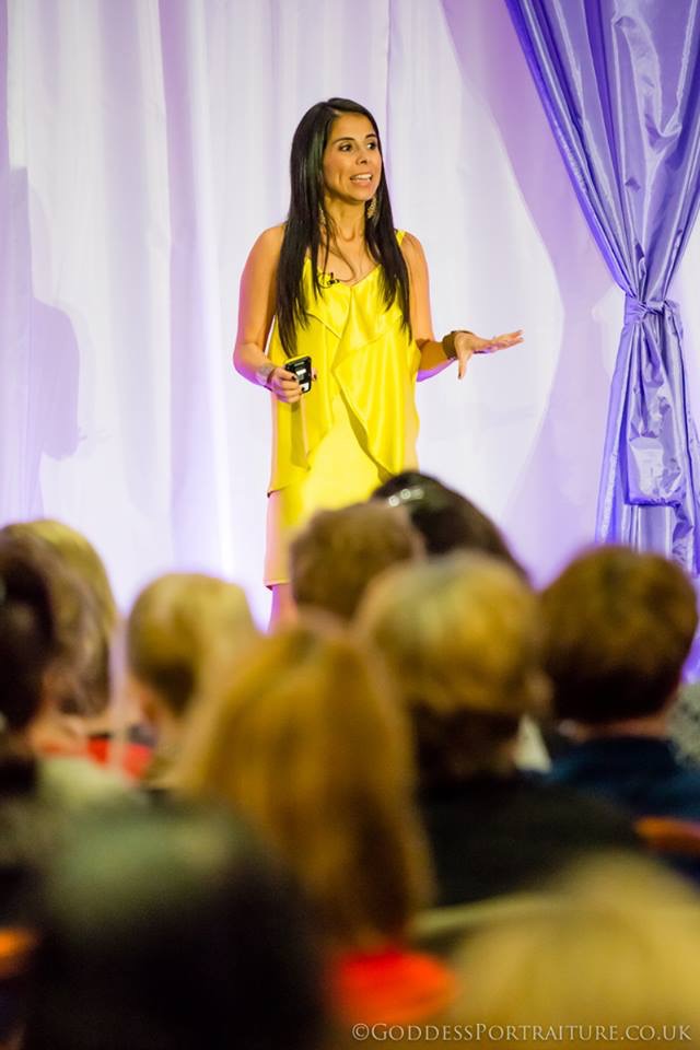 Woman in yellow dress speaking on stage with purple curtains, audience in foreground, holding a microphone.