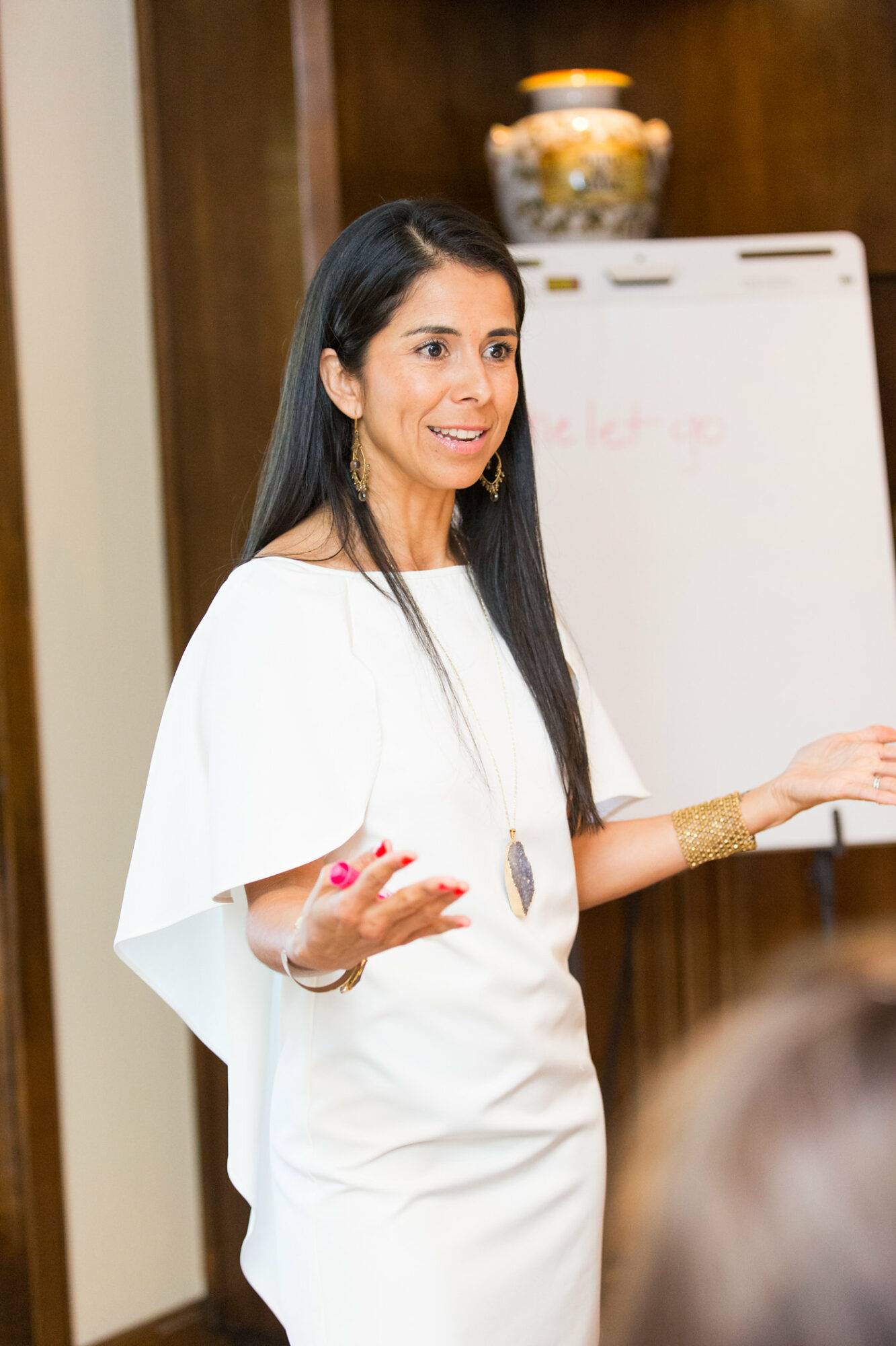 Woman with long dark hair speaking in front of a whiteboard, gesturing with her hands, wearing a white outfit, in a room with wooden walls.