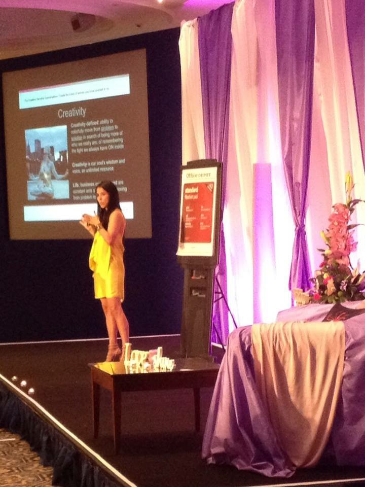 Woman in yellow dress speaking on stage with presentation slide behind her, purple and pink curtains, and a table with flowers and a draped cloth.