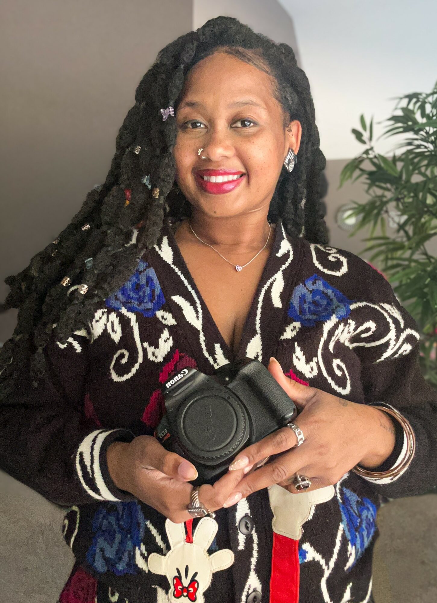 Woman with long dreadlocks holding a camera, smiling, wearing a black and blue patterned top, standing near a plant.