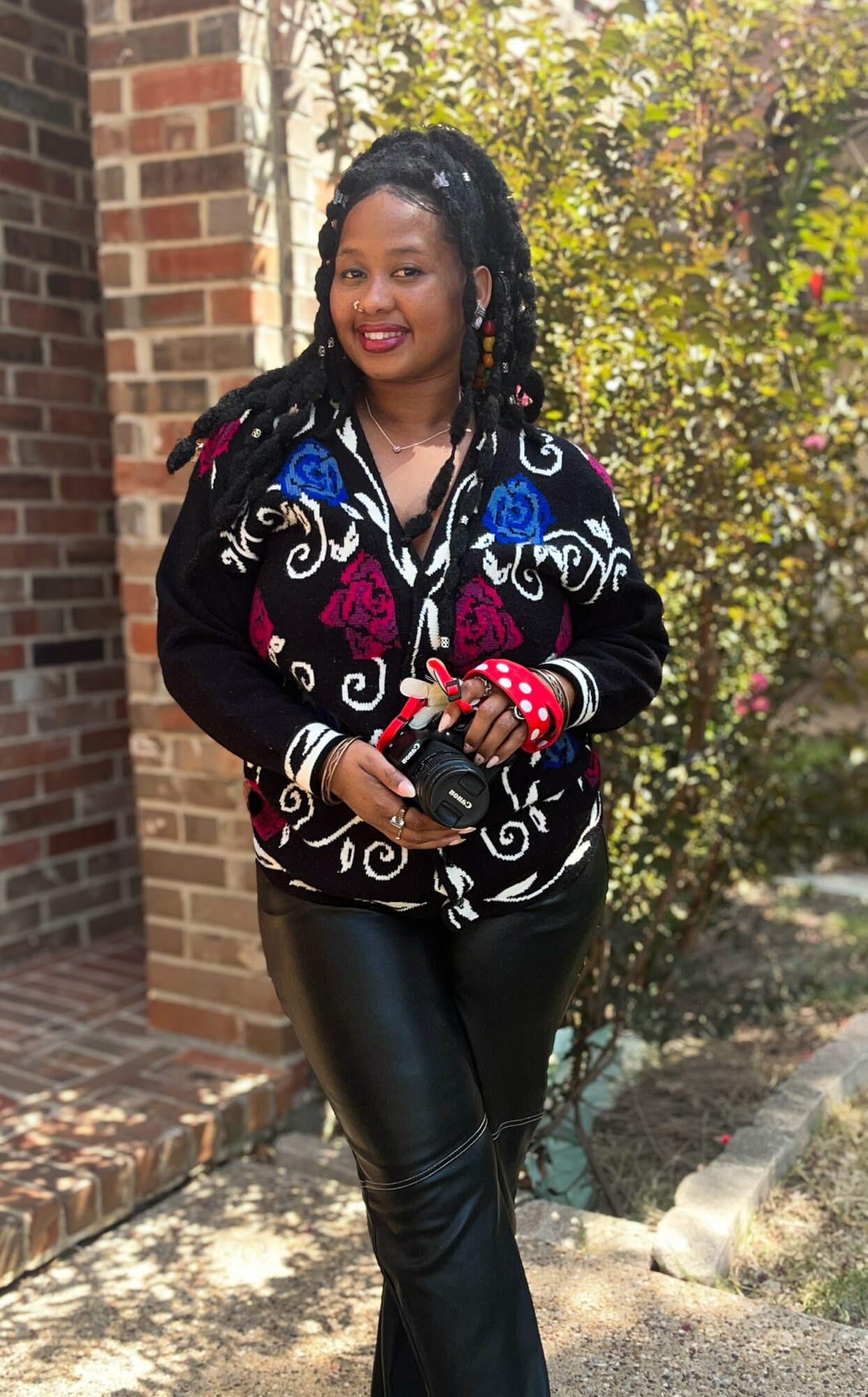 Woman with braided hair smiling outdoors, holding a phone, wearing a colorful jacket and black pants.