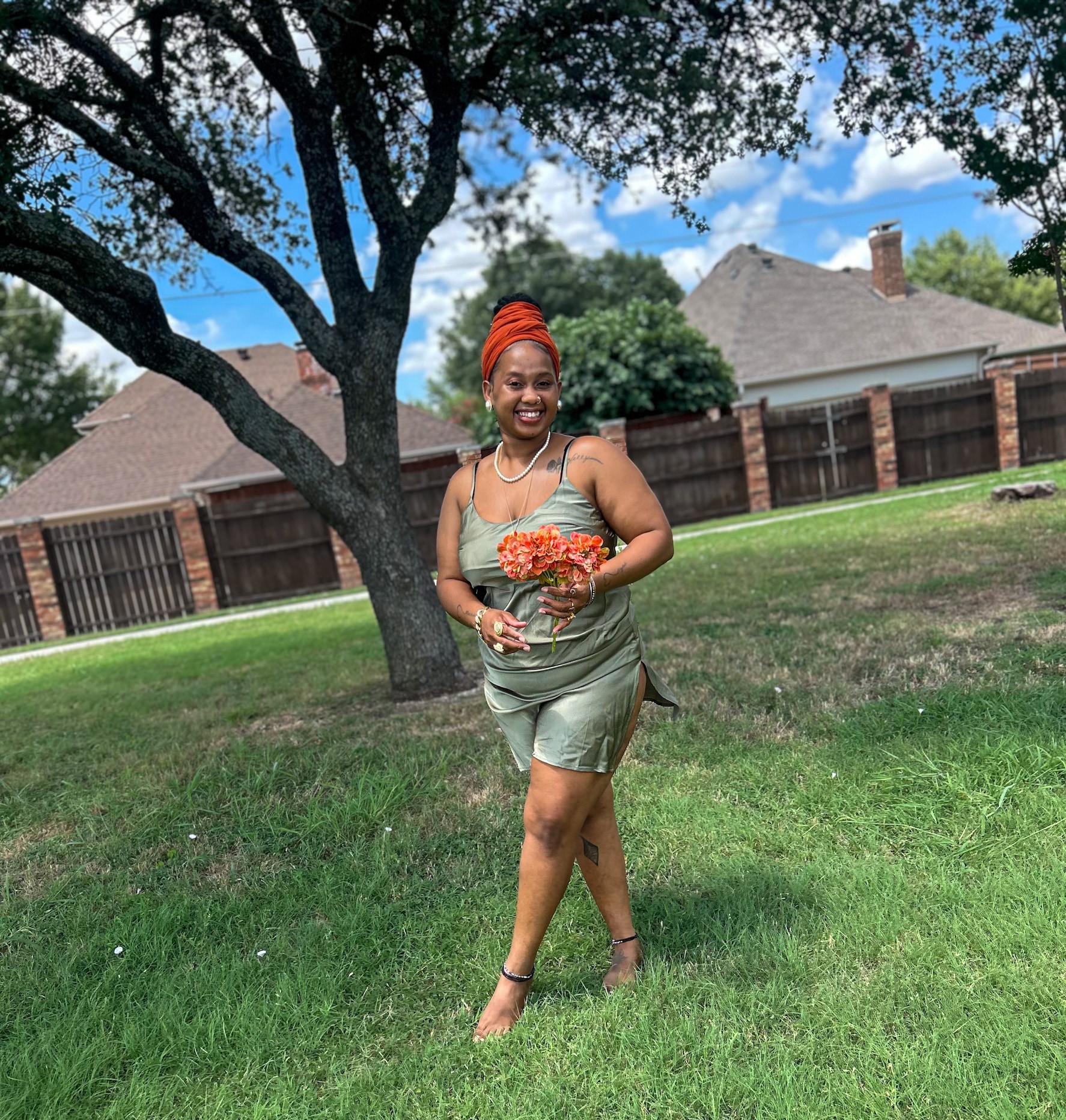 Woman in a green dress holding a bouquet of flowers, smiling, walking on grass near a tree and houses.