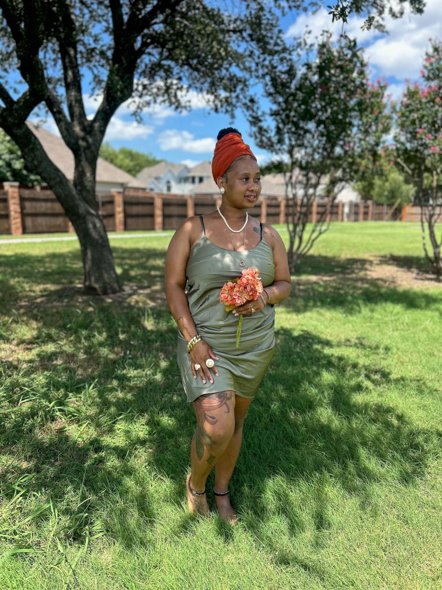 Woman in a green dress holding a bouquet, standing outdoors on grass with trees and a fence in background.