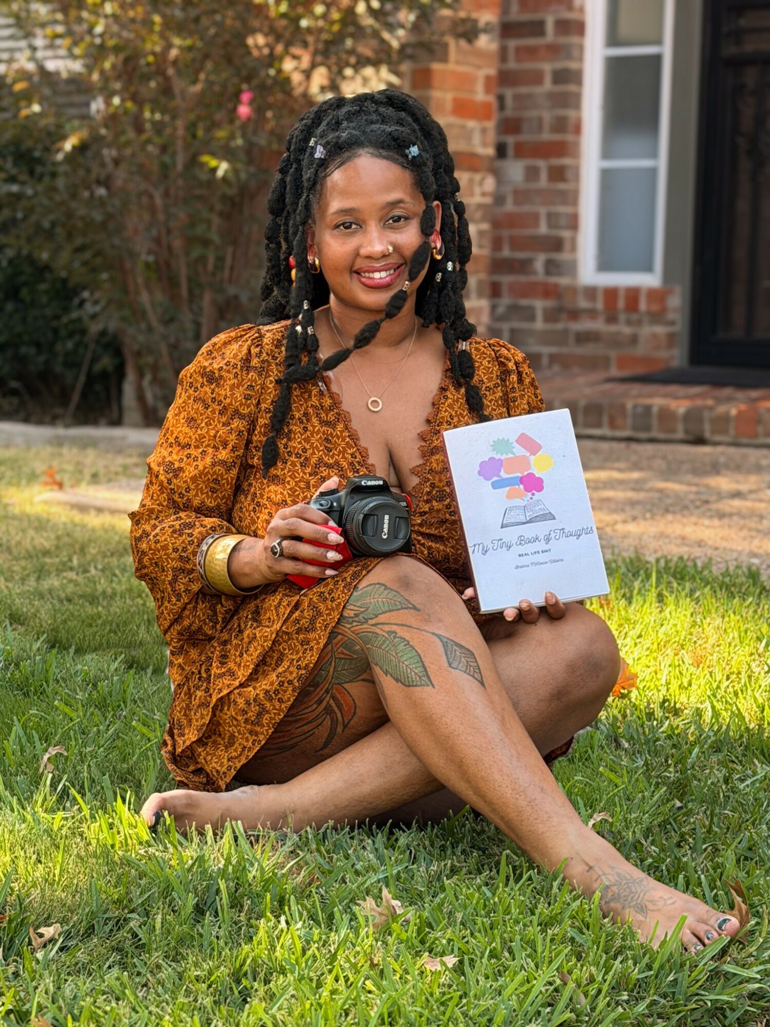 Woman with dreadlocks sitting on grass holding a book and camera outside a brick house, smiling.