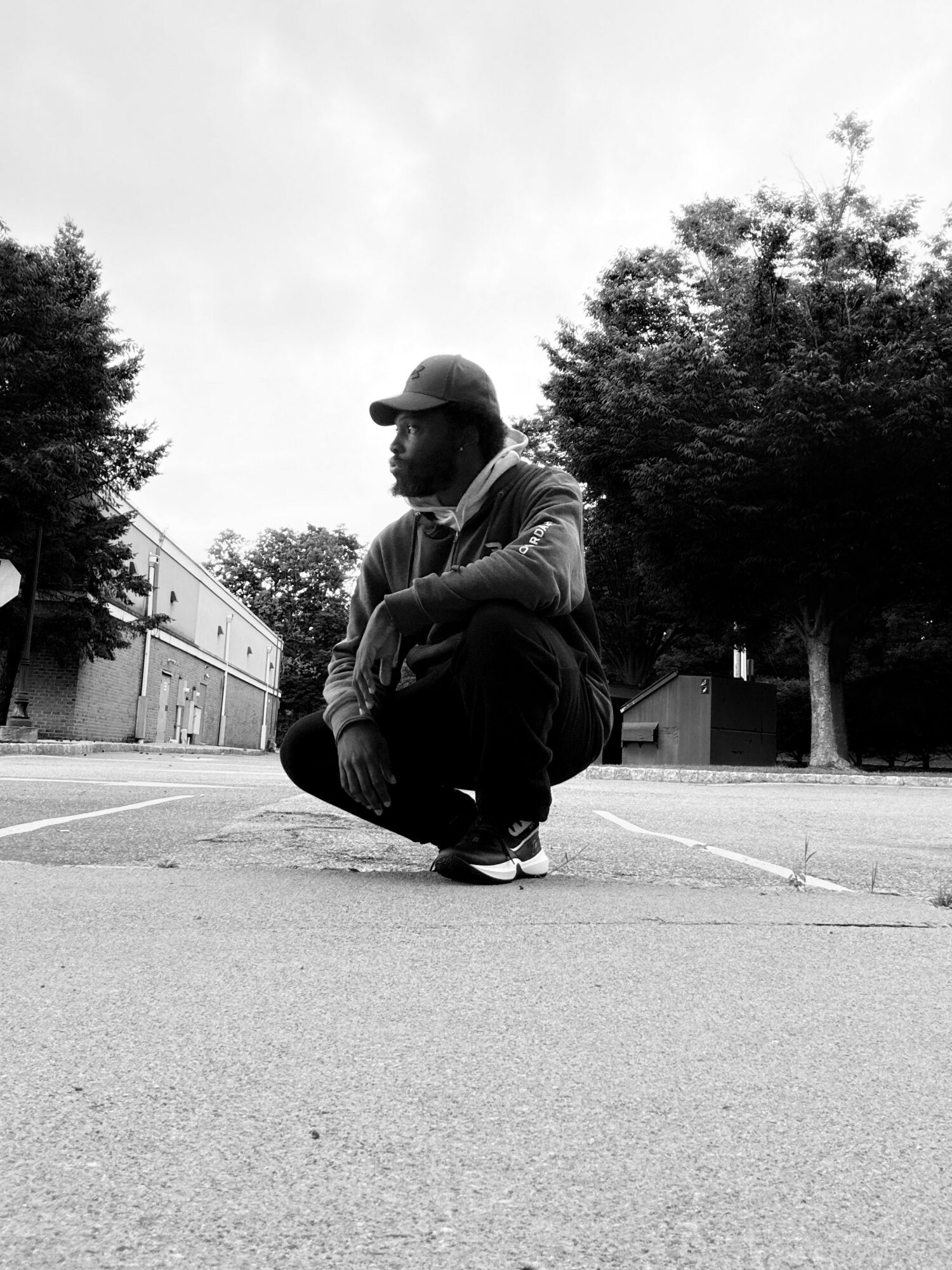Person crouching on pavement with trees and buildings in background, overcast sky, black and white photo.