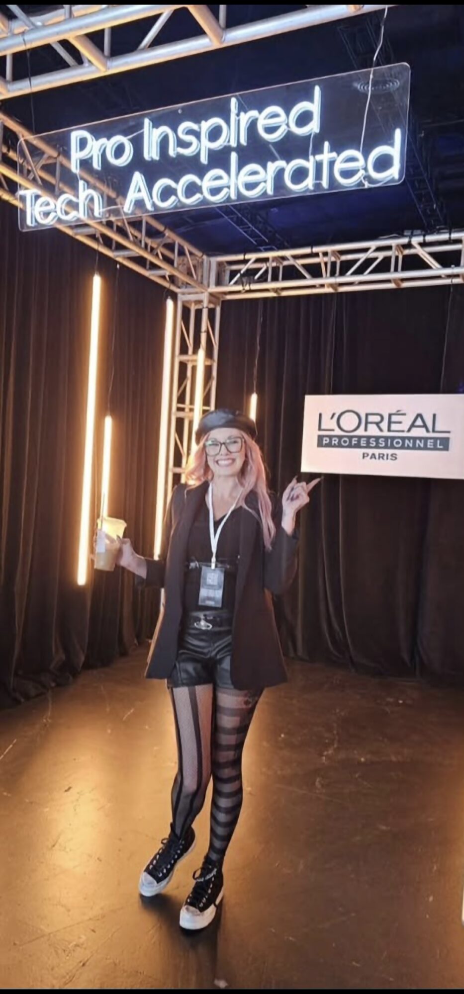 Person standing on stage with a sign and neon sign above, holding a microphone, smiling, wearing striped tights and glasses.