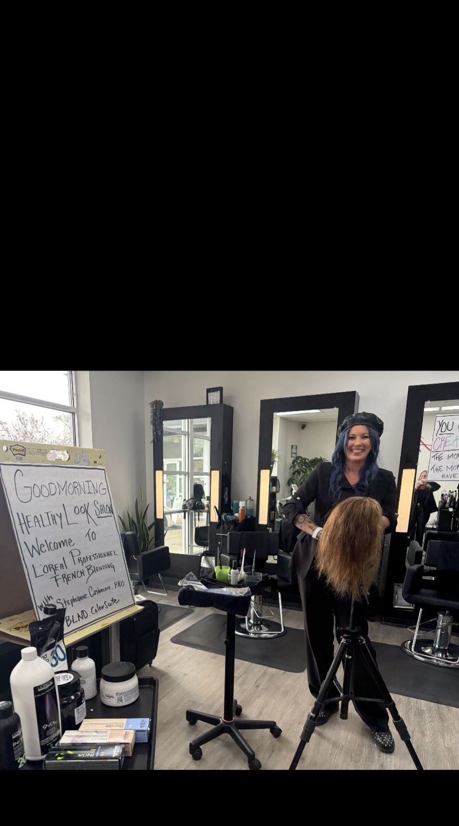 Woman with long dark hair smiling in a hair salon, facing a mirror with hair styling tools and products around.