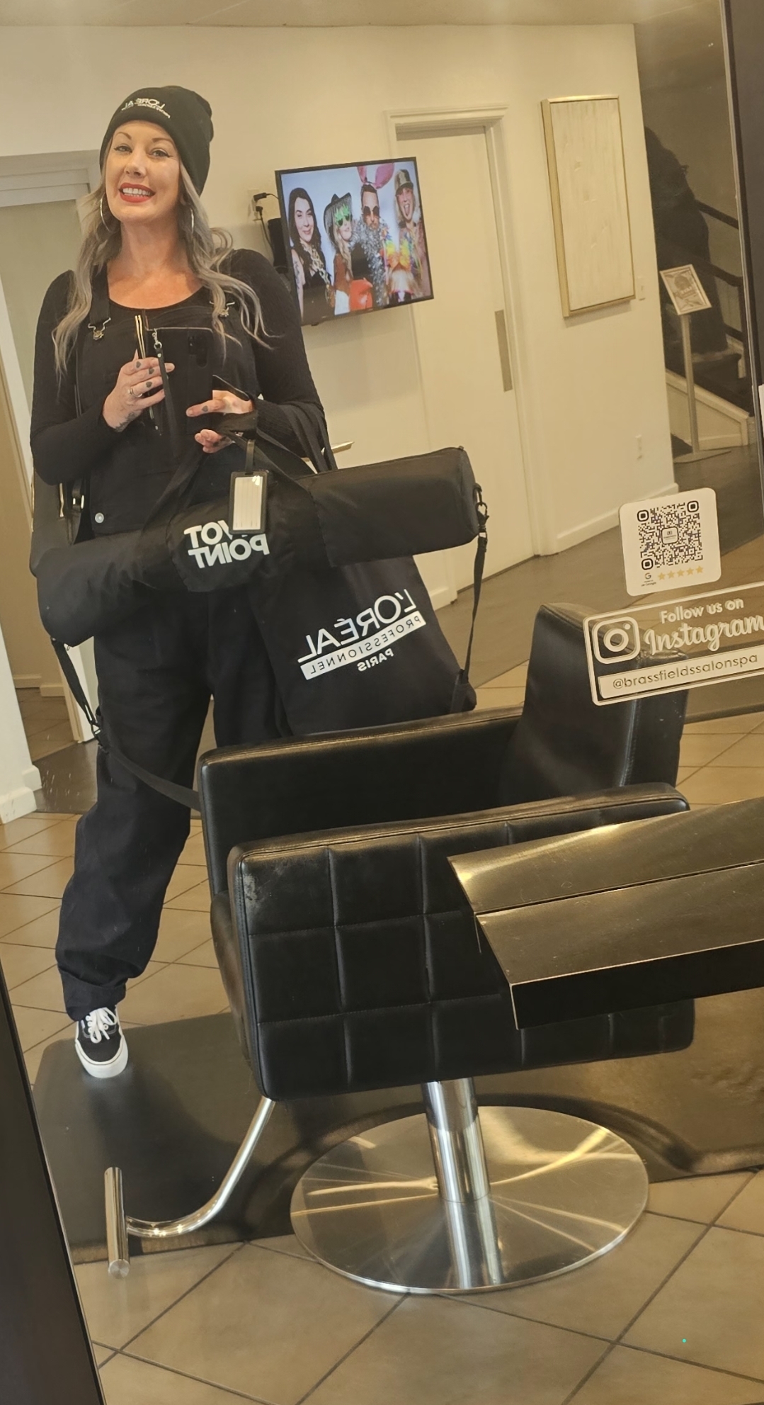 Woman taking a selfie in a salon, holding a phone, with salon chairs and a TV in the background.