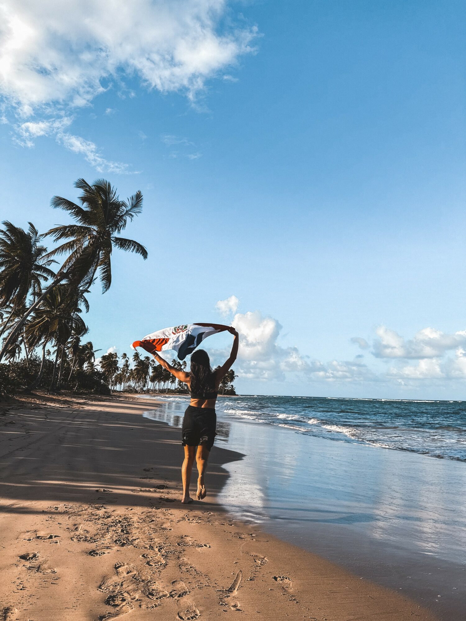 Person holding a surfboard on a sandy beach with palm trees and ocean, under a partly cloudy sky.
