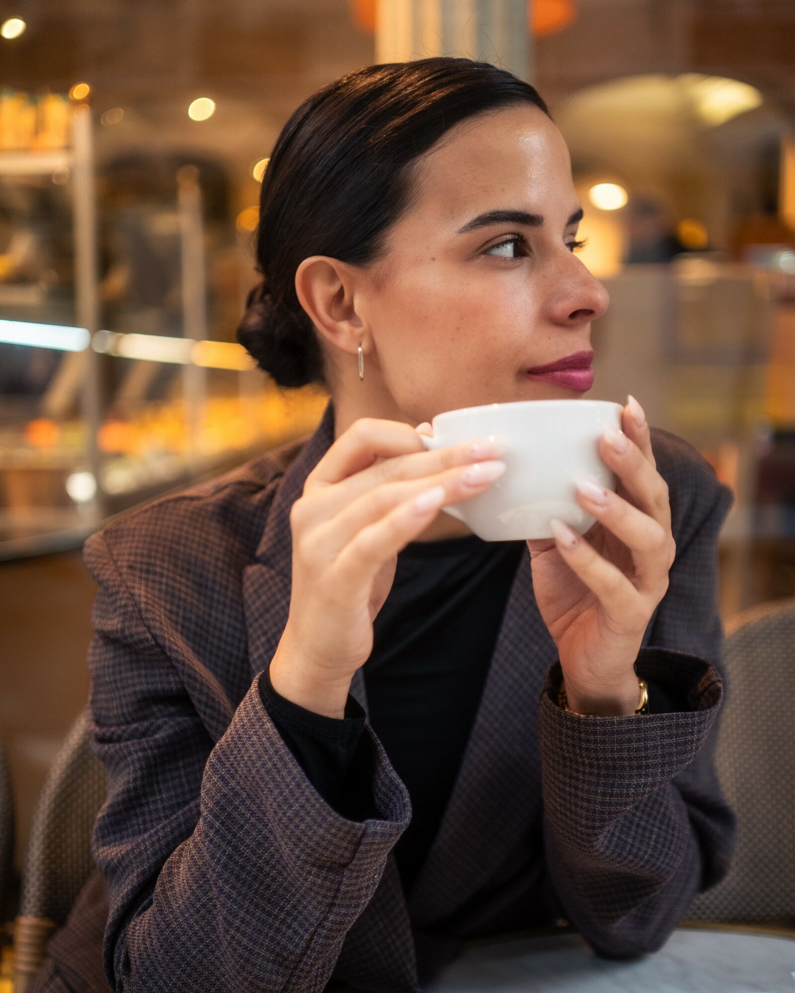 Woman with dark hair in a bun holding a white cup, sitting indoors with blurred background lights.