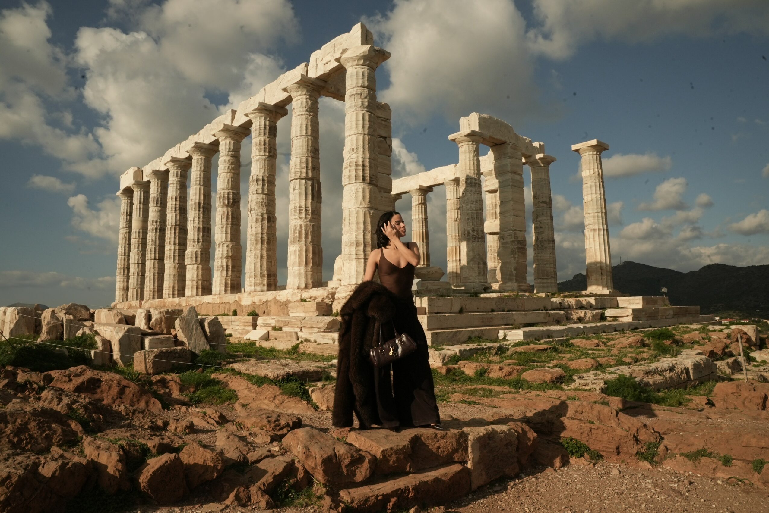 Woman in black dress standing near ancient Greek ruins with columns, under a partly cloudy sky.