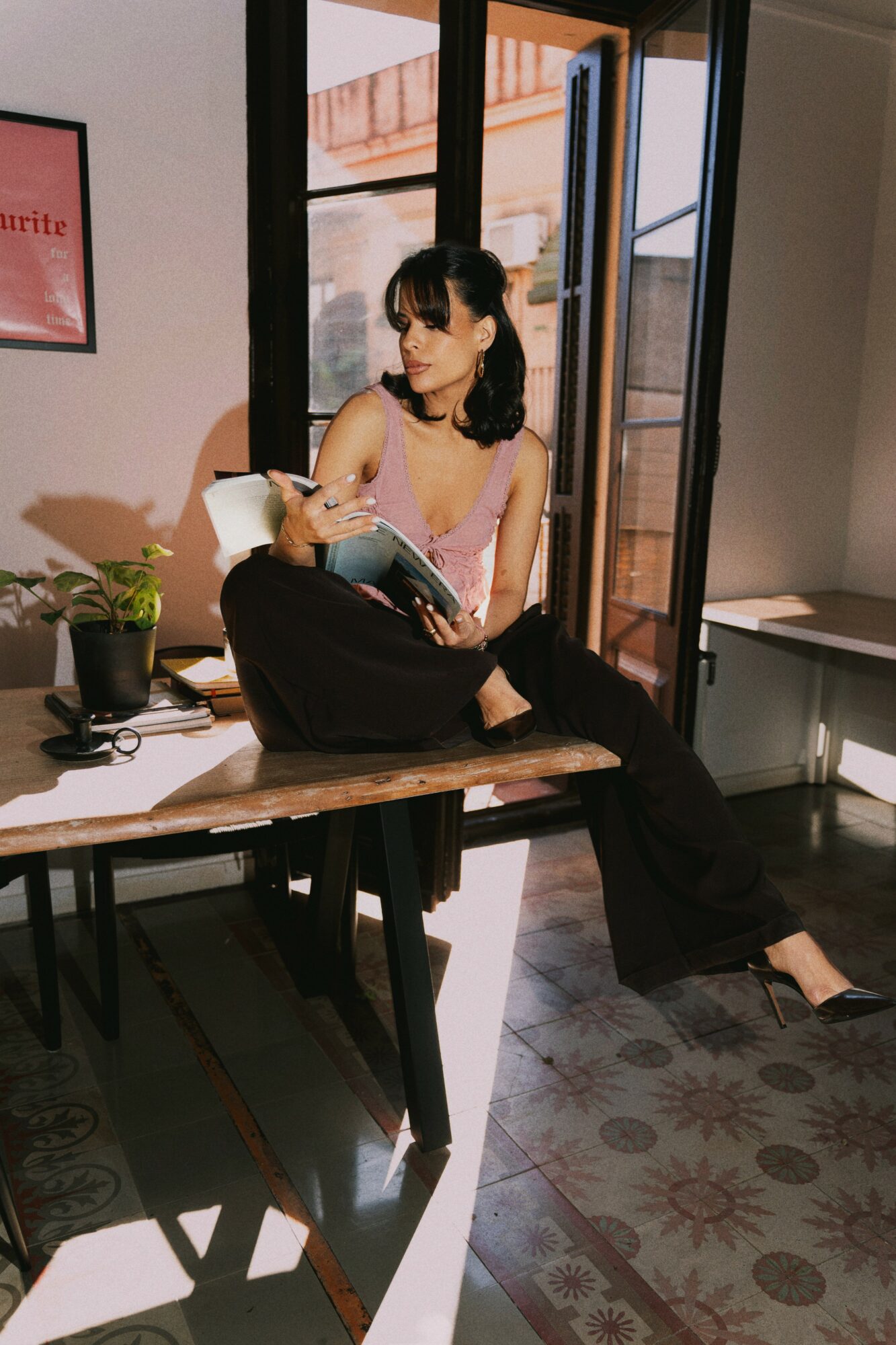 Woman sitting on a desk reading a book, with sunlight streaming through open windows behind her.