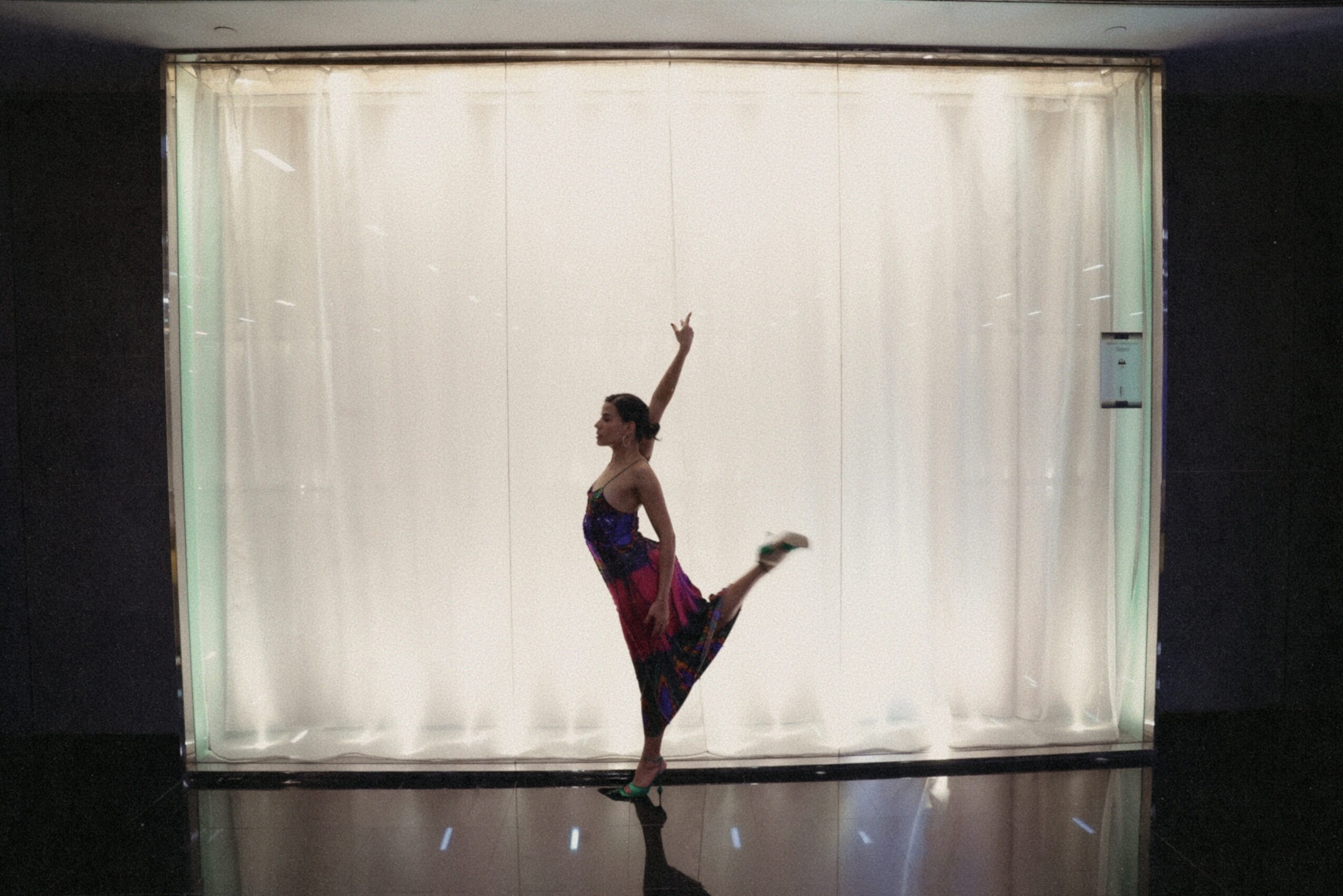 A person in a dance pose with one arm raised and one leg lifted, standing in front of a lit curtain.