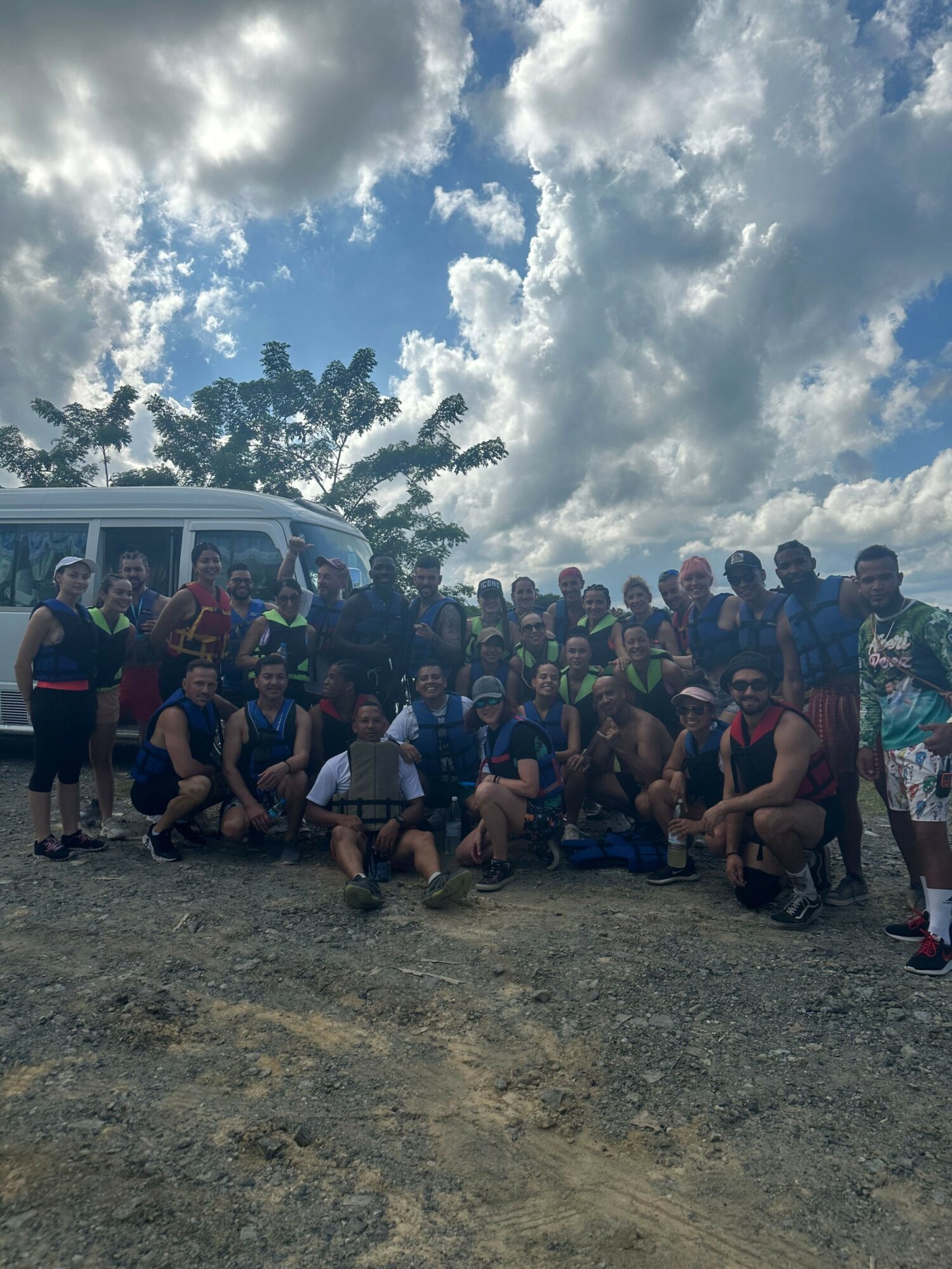 Group of people outdoors on a dirt ground with trees and cloudy sky in the background.