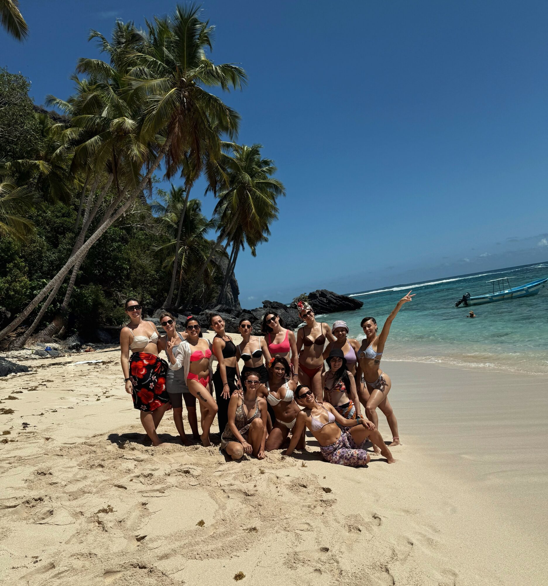 Group of people on a sandy beach with palm trees and ocean, some standing and some sitting, sunny day.