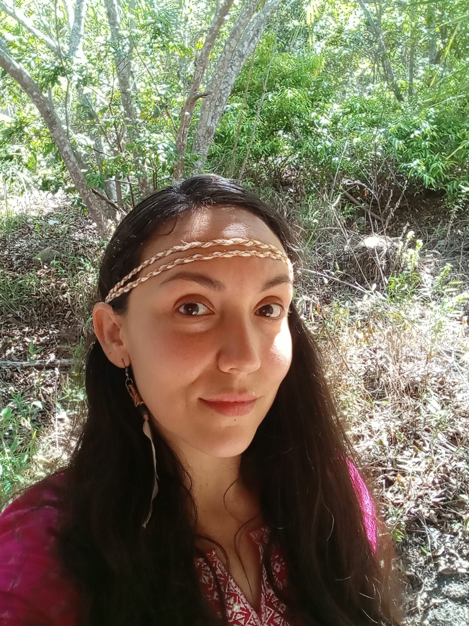 Young woman with long dark hair and jewelry, outdoors among trees and greenery.