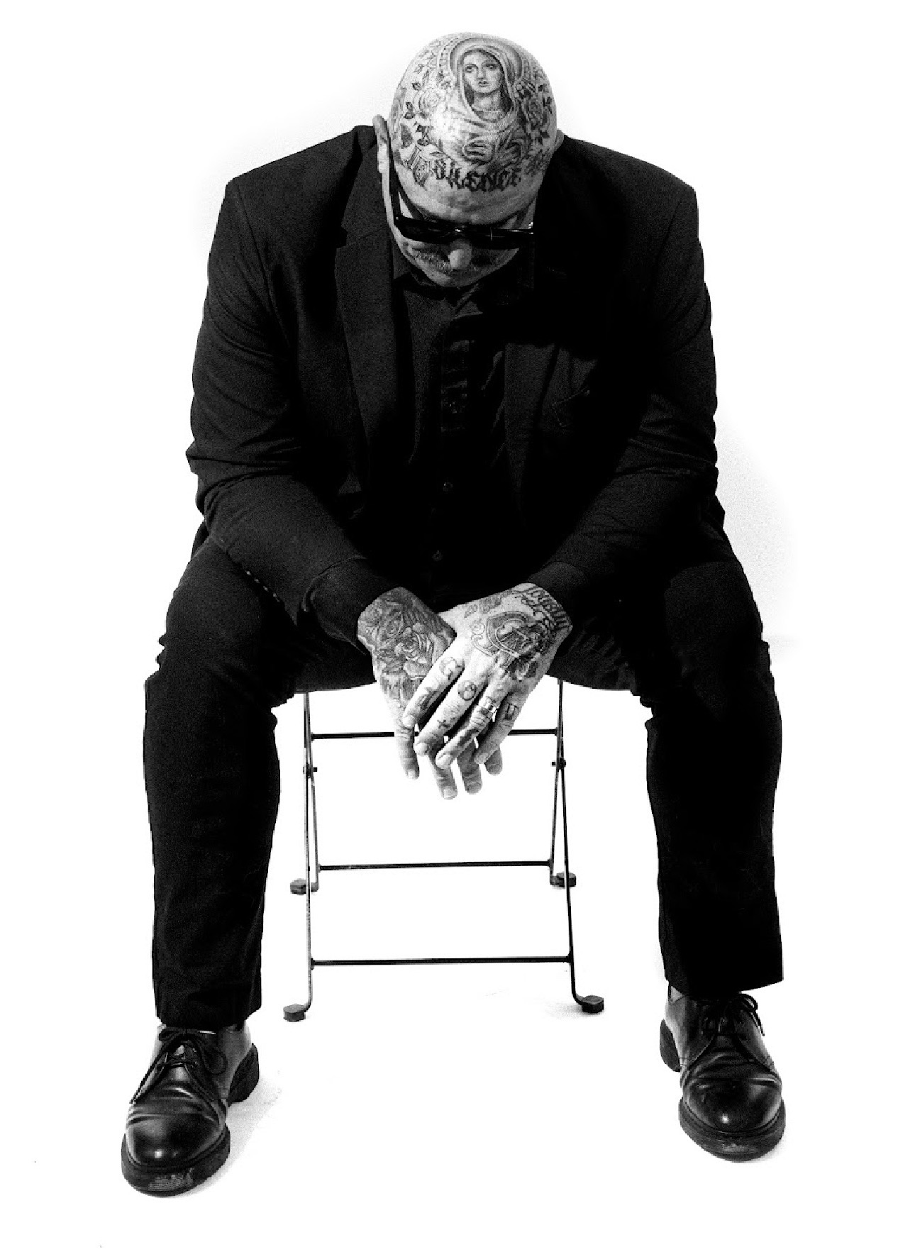 Person with tattoos and a bandana sitting on a small stool, head bowed, in black clothing, against a white background.