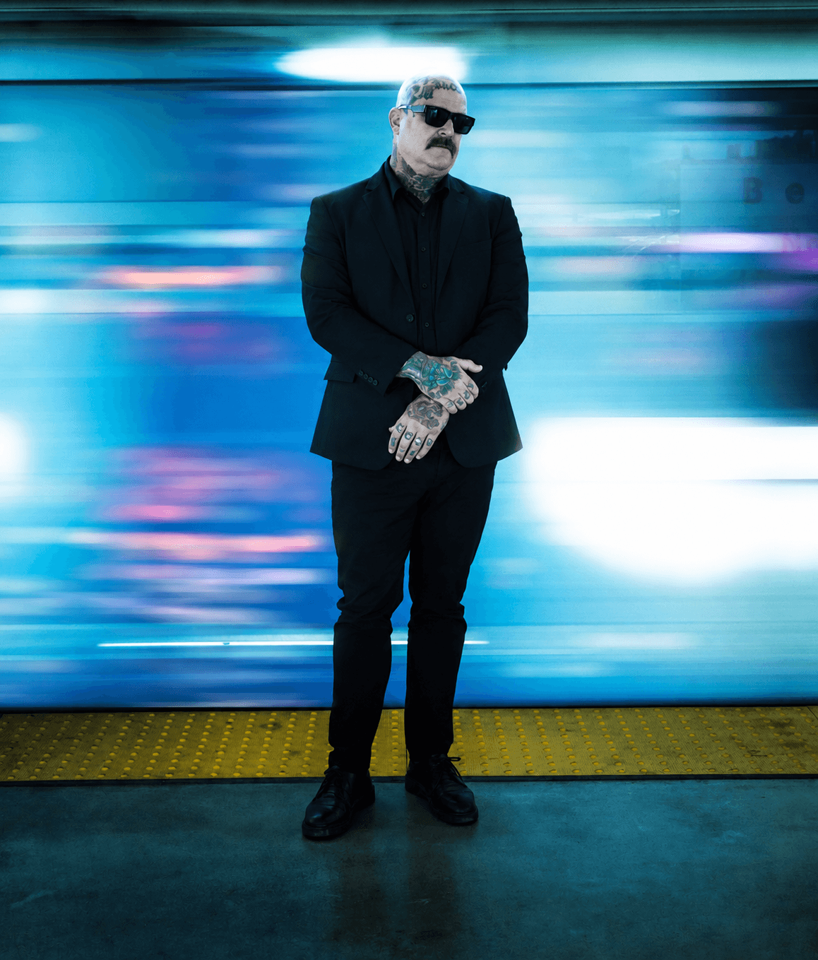 Man with sunglasses and tattoos stands with arms crossed in front of a colorful, blurred background at a station or platform.