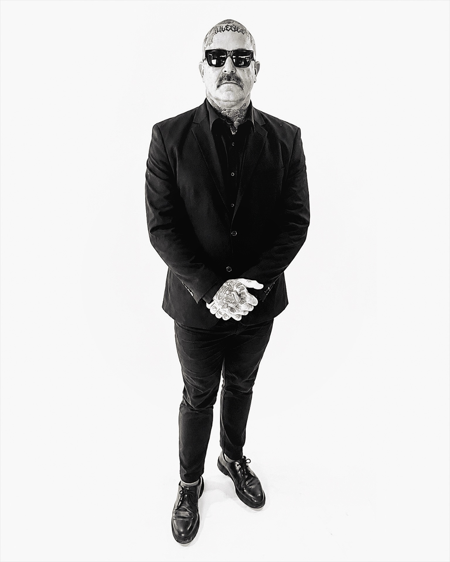 Man with sunglasses, beard, and mustache wearing a black suit and gloves, standing with hands clasped, facing forward.