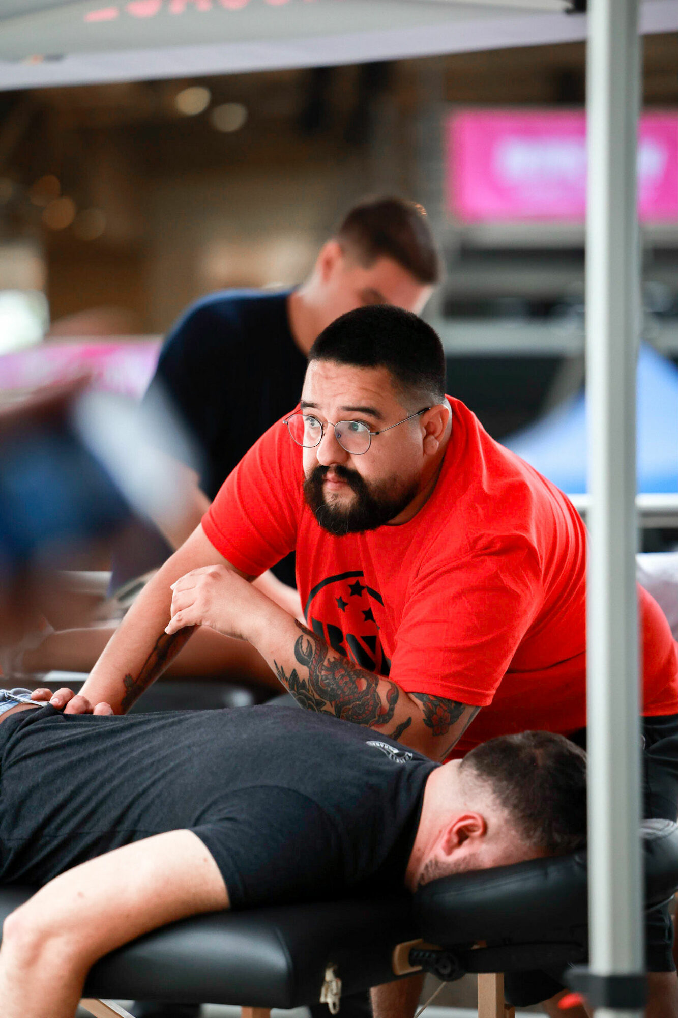Man with a beard and tattoos lying face down on a massage table, wearing a red shirt, in a wellness setting.