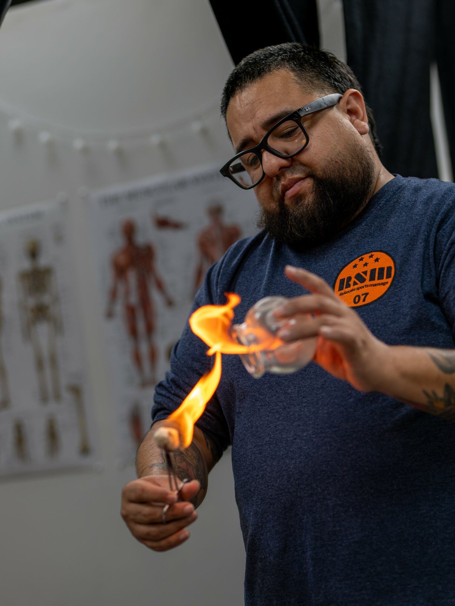 Man with glasses holding a flaming object in a laboratory setting, with anatomical posters in the background.