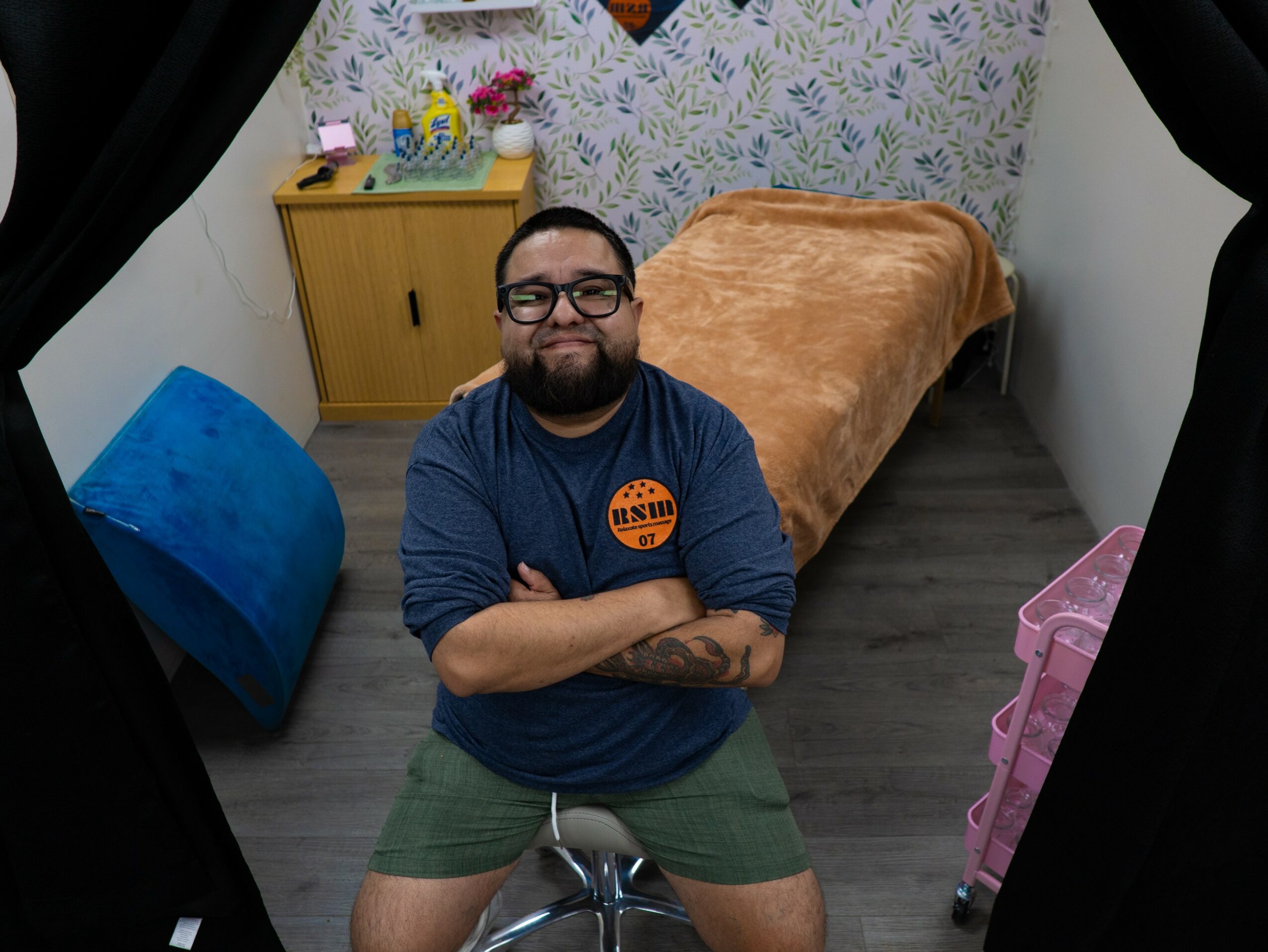 Person with glasses and beard smiling, arms crossed, sitting on a stool in a room with a bed and colorful decor.