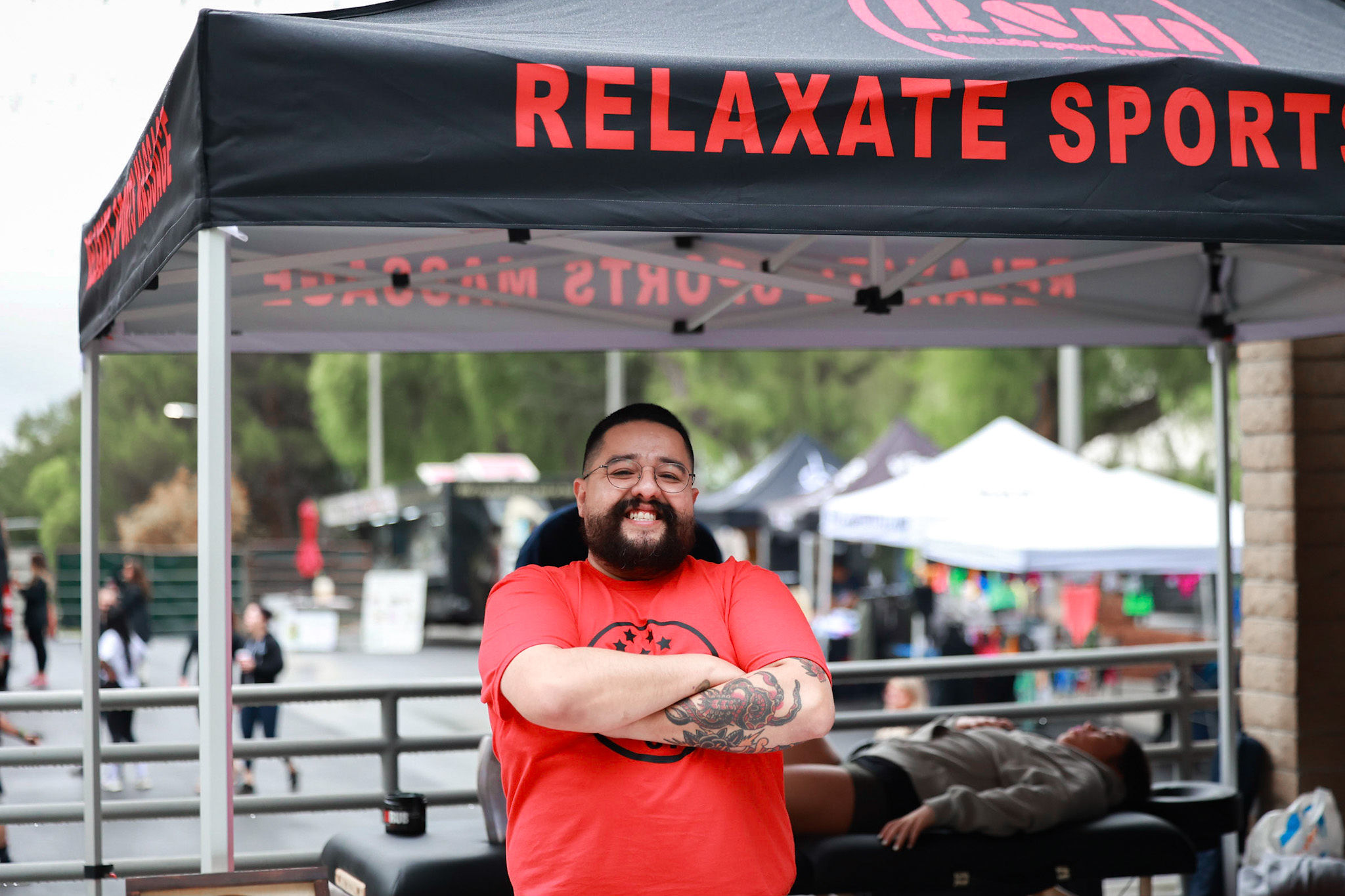 Smiling man with a beard and tattoos, wearing a red shirt, standing under a canopy with 'RELAXATE SPORT' text.